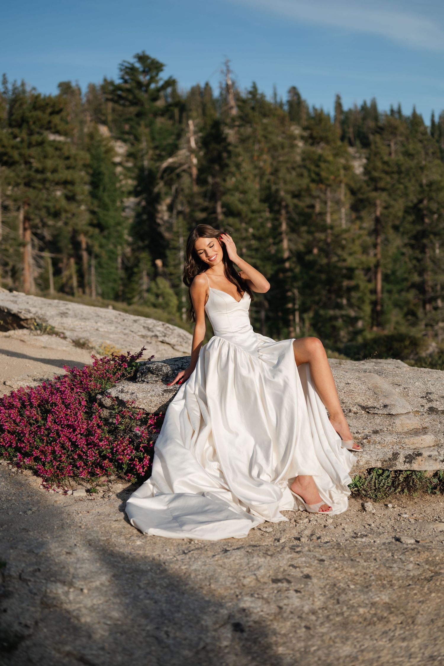 Bride in white satin gown sits on rocky terrain with pink wildflowers and pine forest backdrop in golden light.