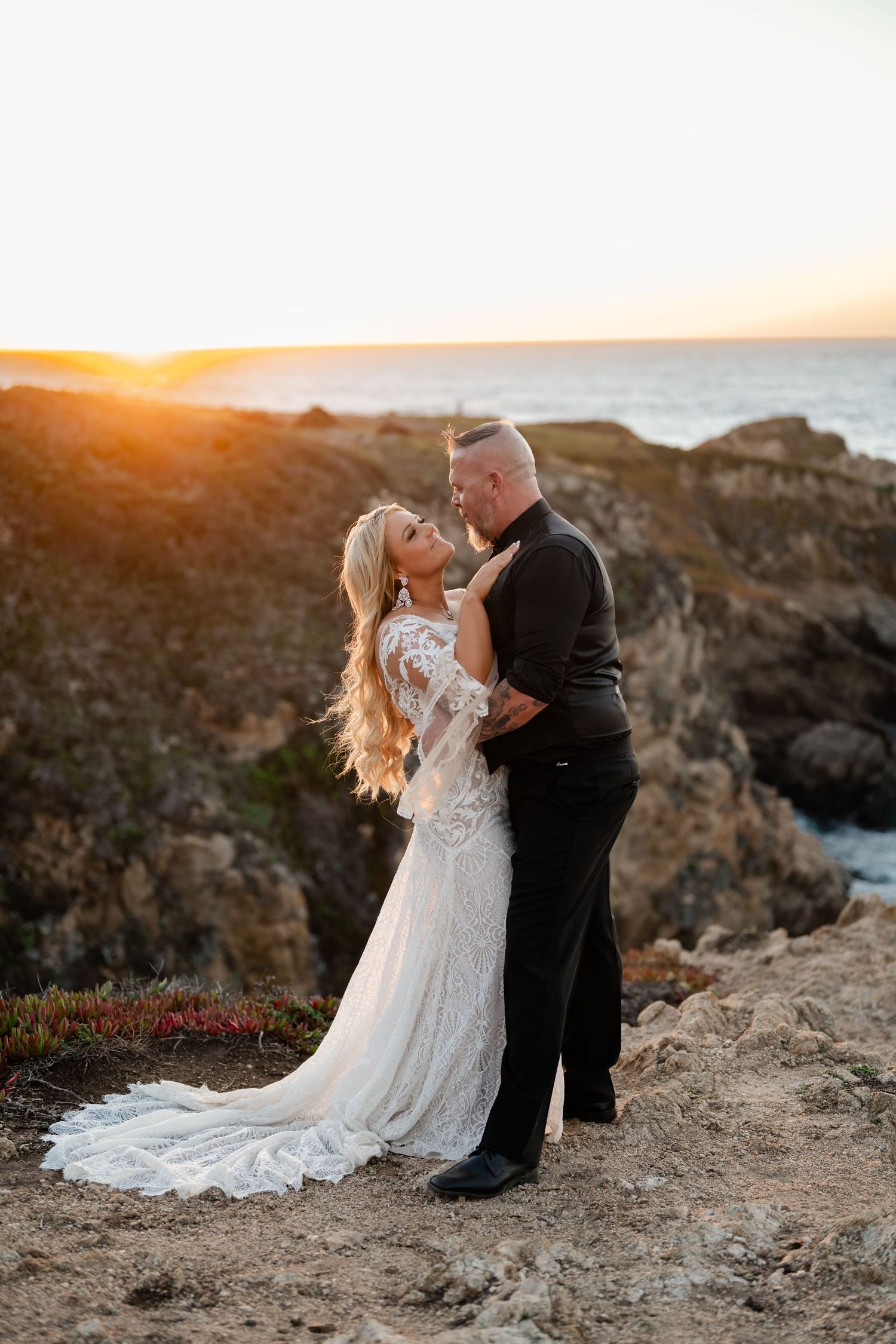 Wedding couple poses dramatically on rocky oceanside cliffs at sunset.