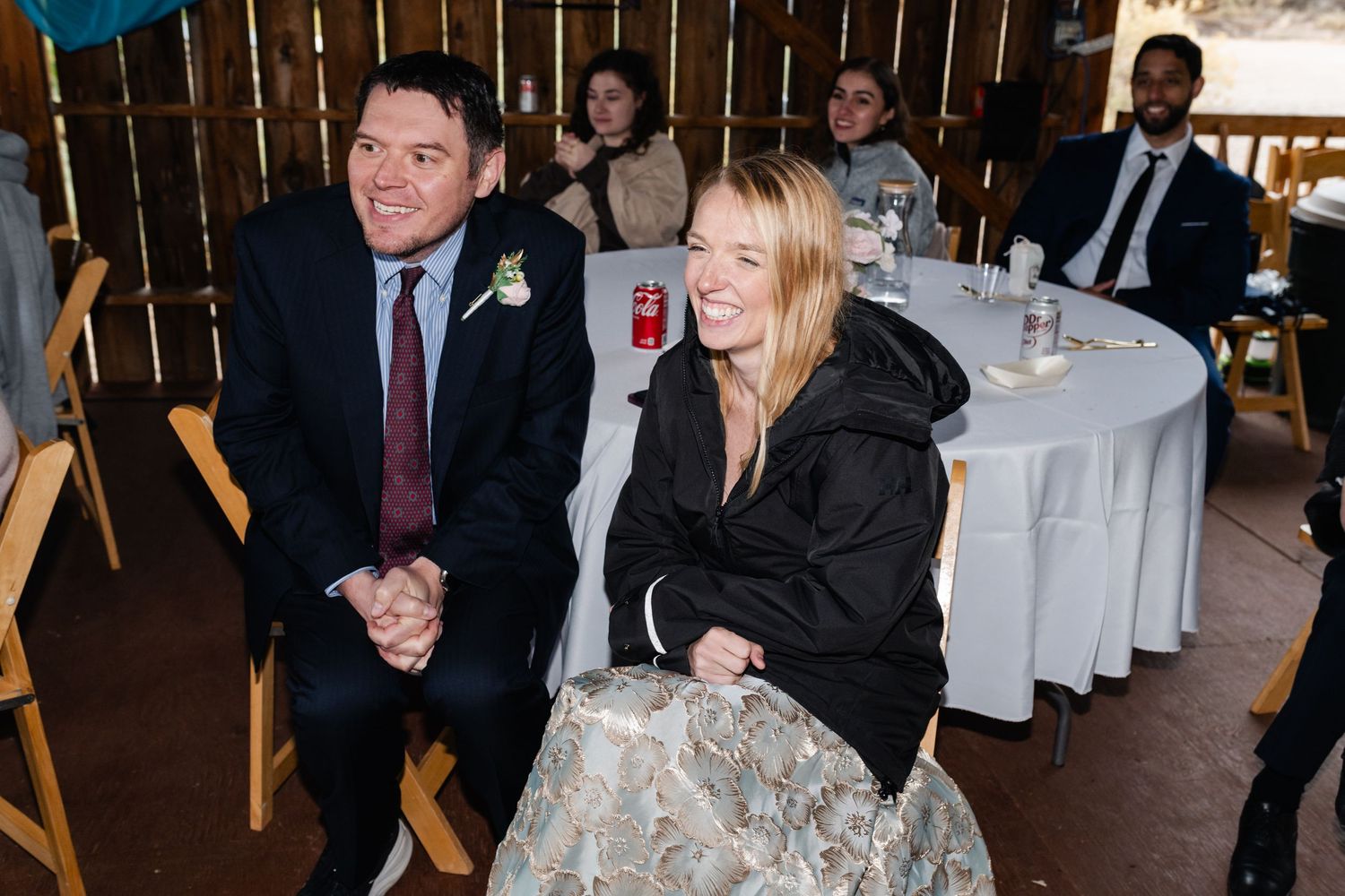 Wedding guests gather in a rustic venue decorated with string lights and wooden beams.