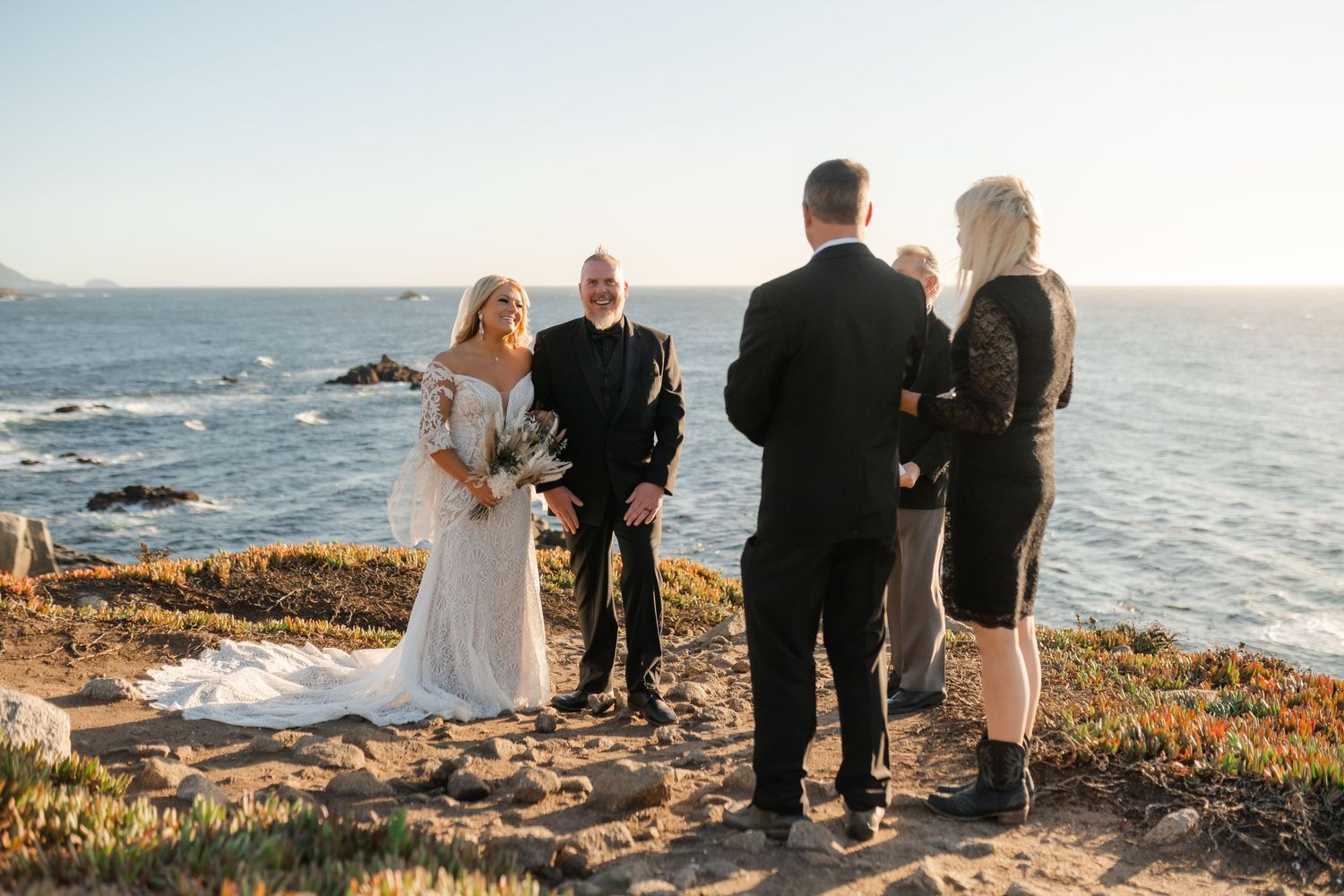 An intimate wedding ceremony unfolds against a backdrop of ocean waves and rocky coastline.