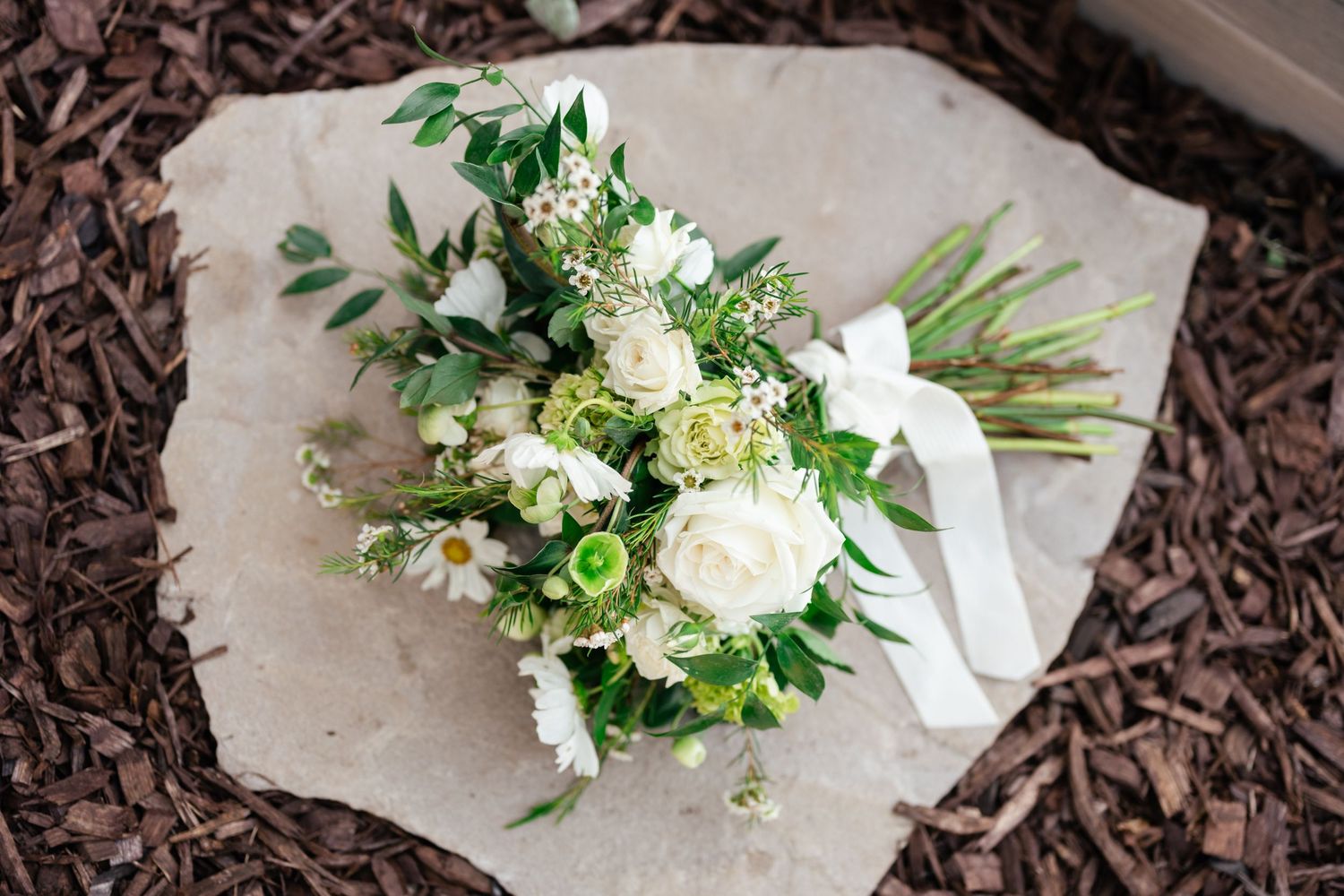 Elegant white and green wedding bouquet with roses and mixed flowers wrapped in light fabric lying on mulch.