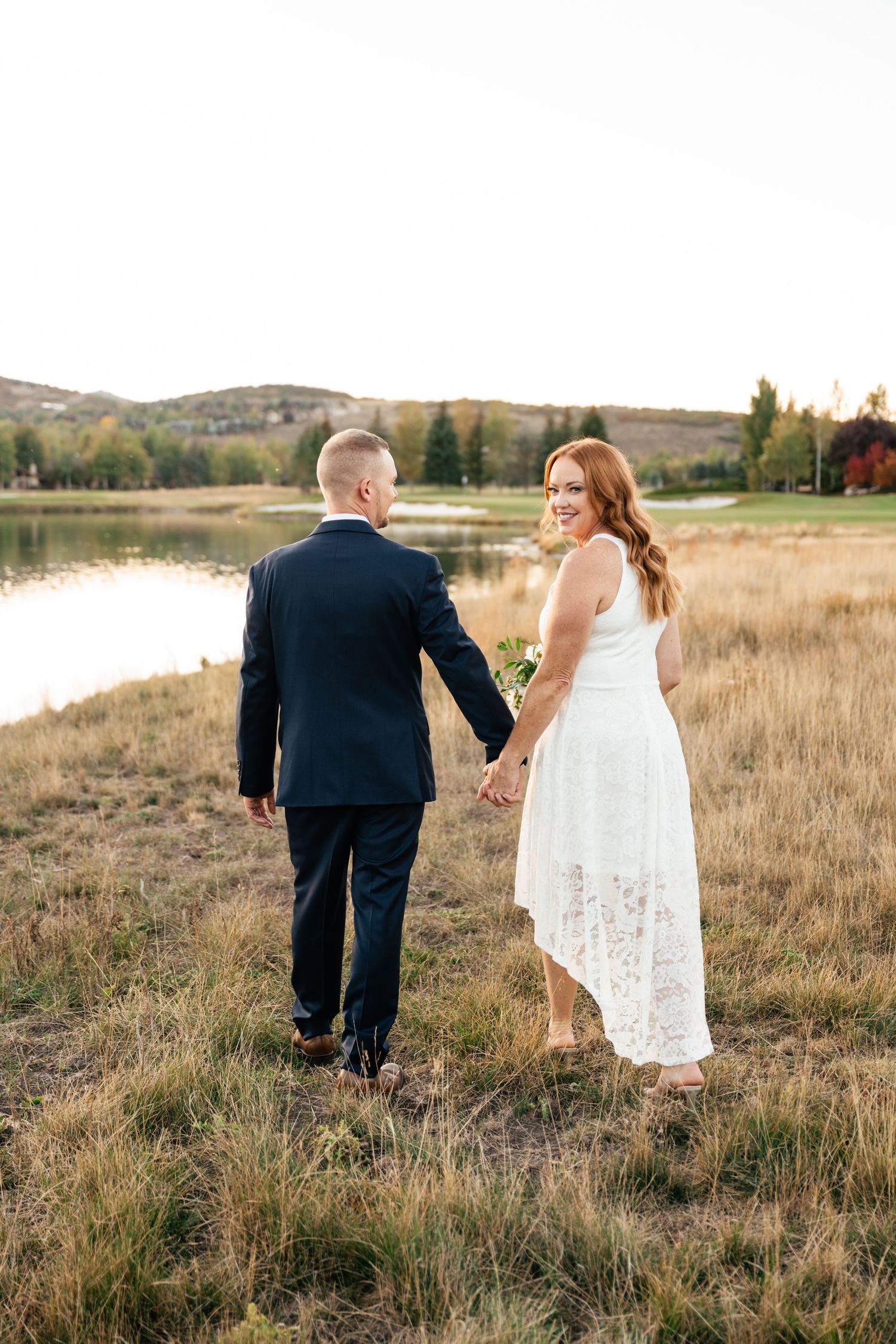 A couple walks hand in hand through a grassy field near a lake with mountains in the background at sunset.