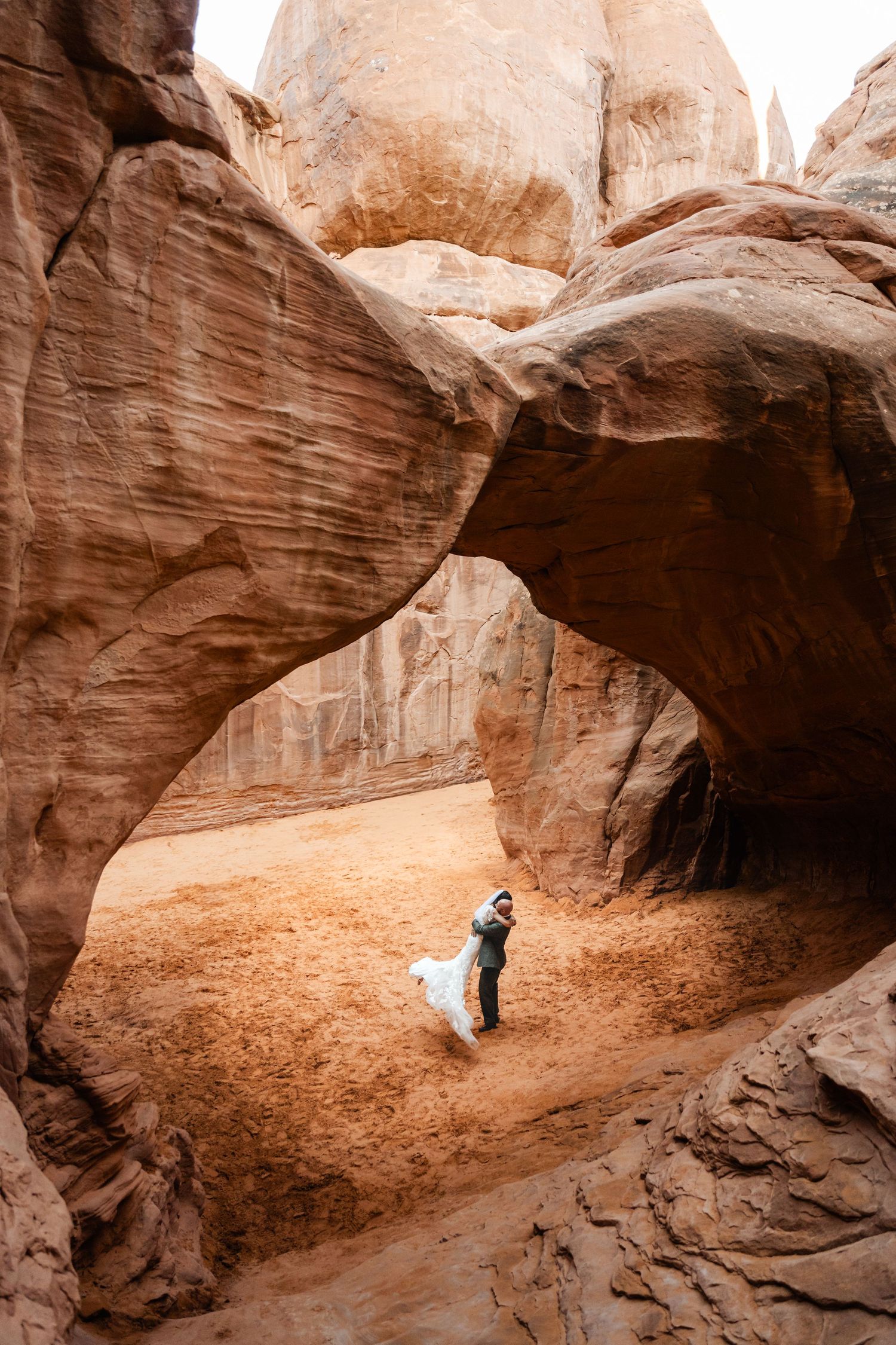 A wedding couple embraces under a massive natural rock arch in the desert.
