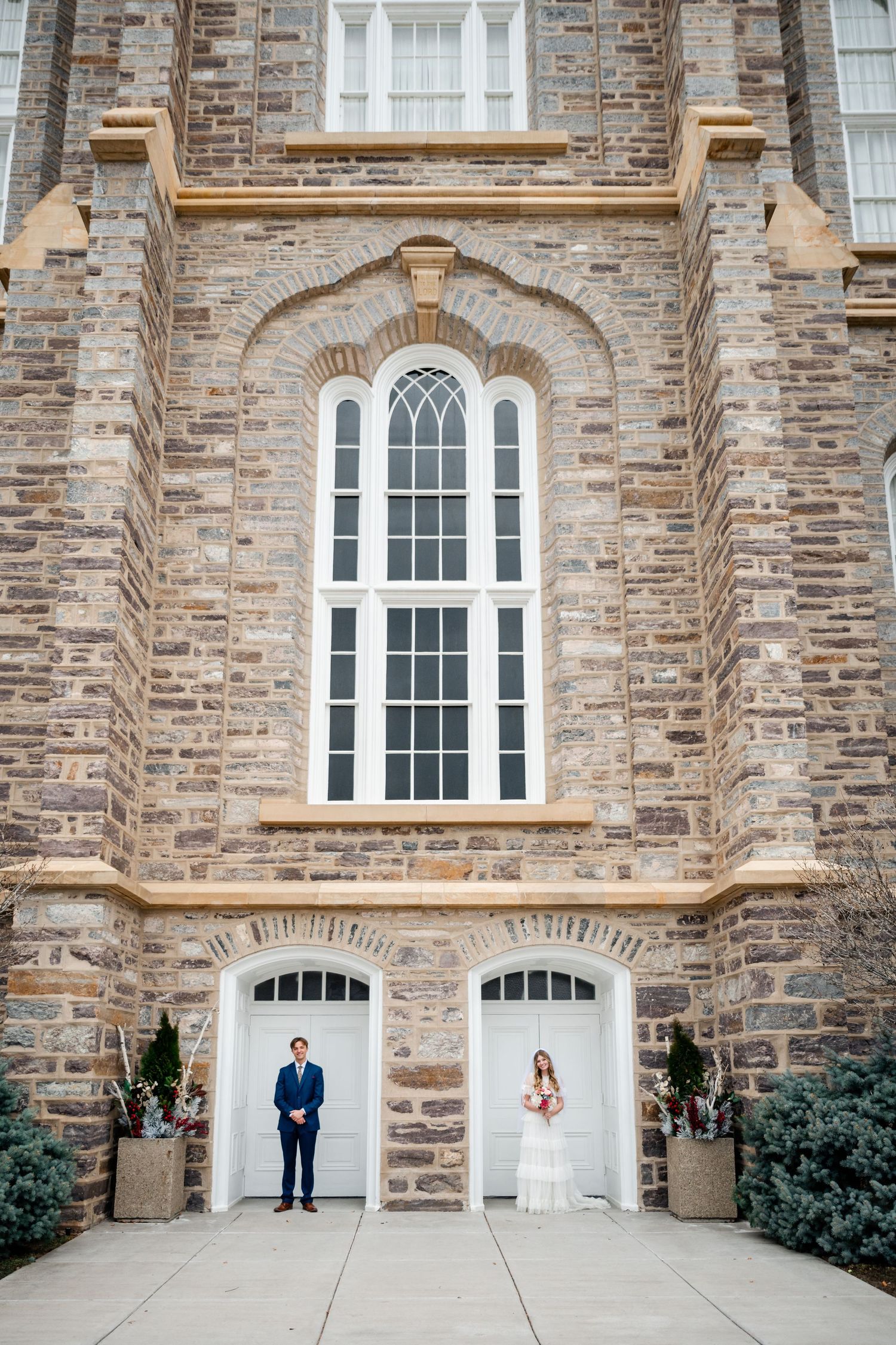 A couple stands in separate white archways beneath a tall stone church window with ornate architectural details.