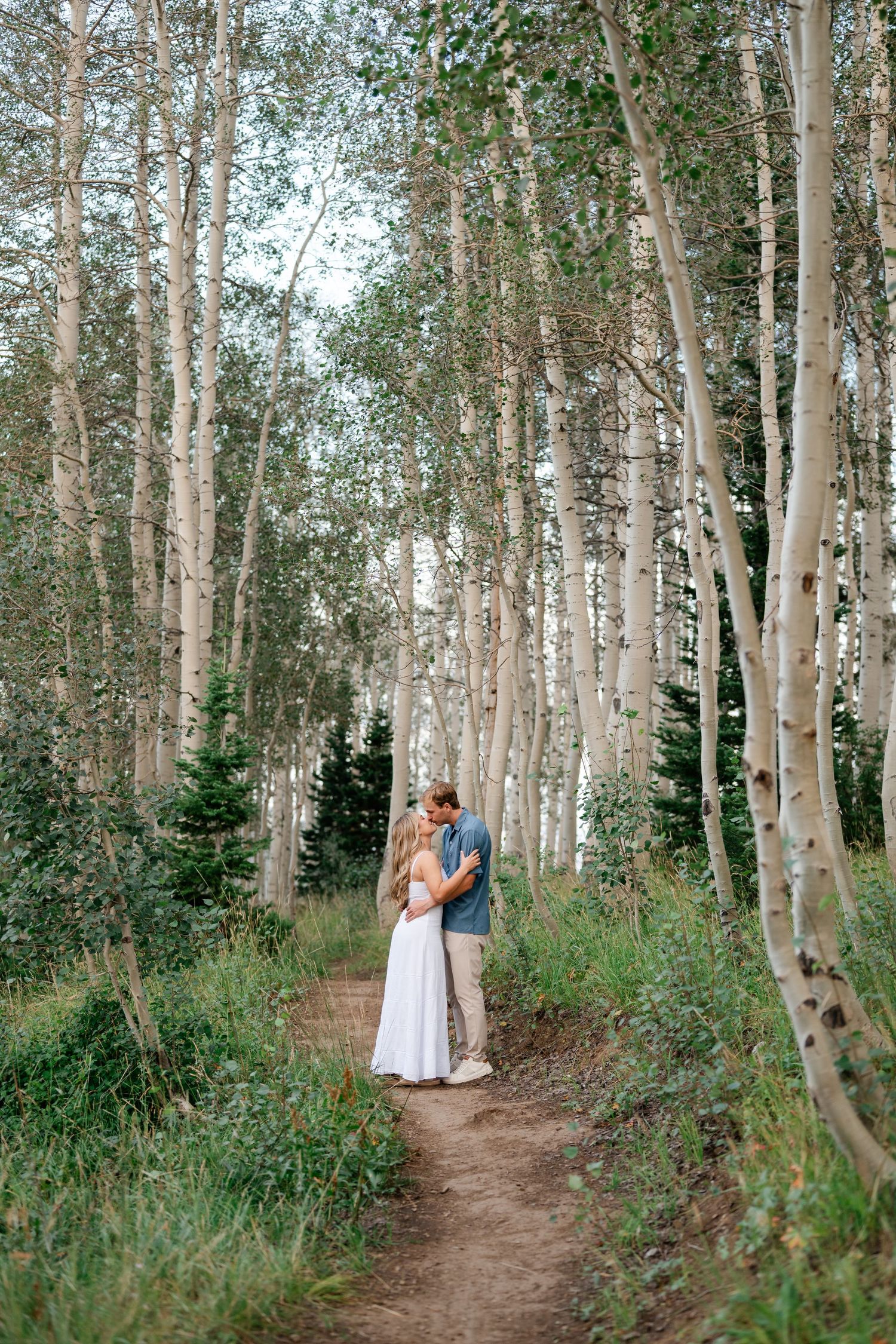 Couple sharing a romantic kiss on a forest path surrounded by tall white birch trees and lush greenery above Park City.