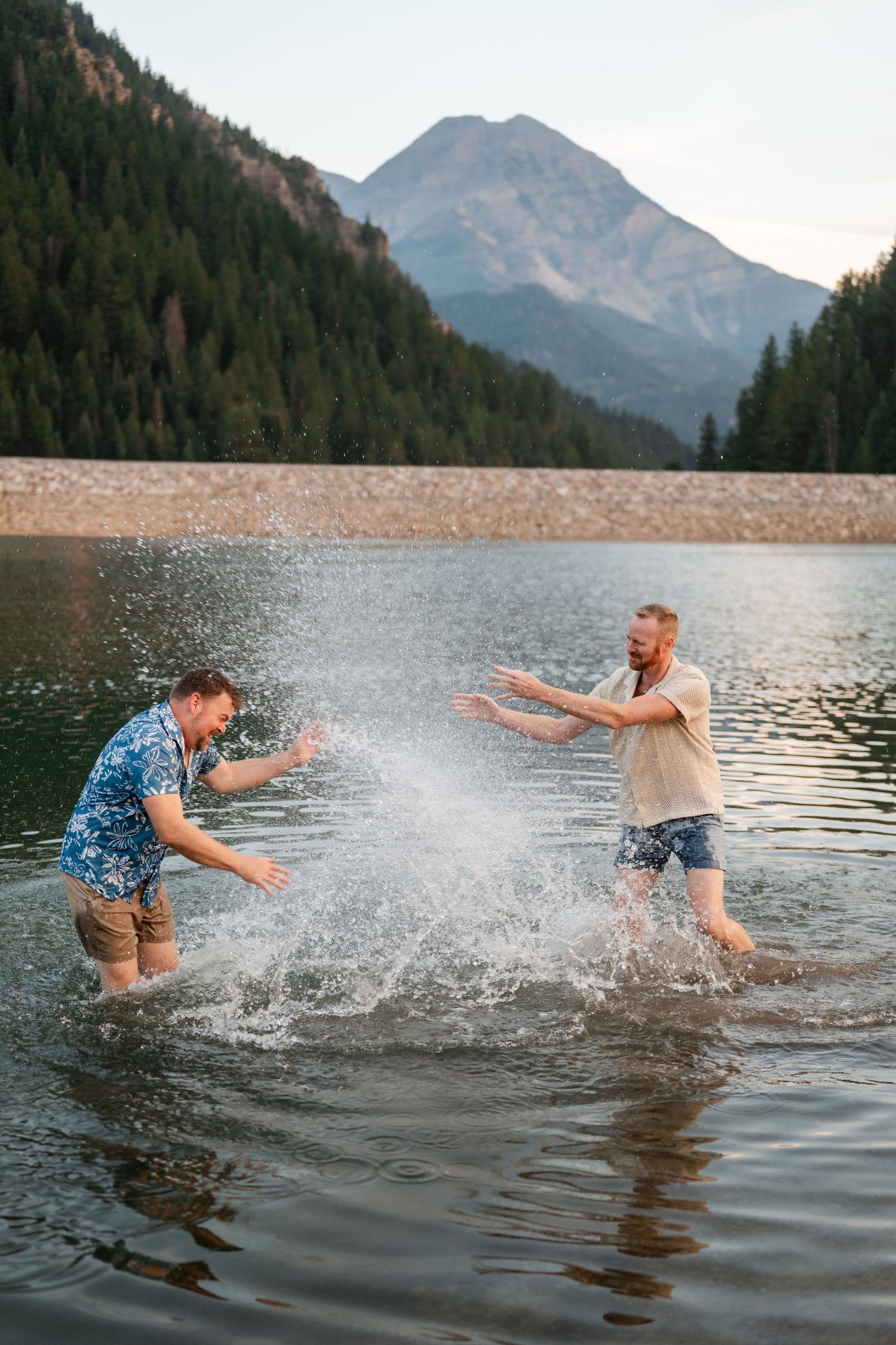 Couple having a water fight at Tibble Fork. Two men splashing water in a mountain lake surrounded by pine forests and rocky peaks at dusk.