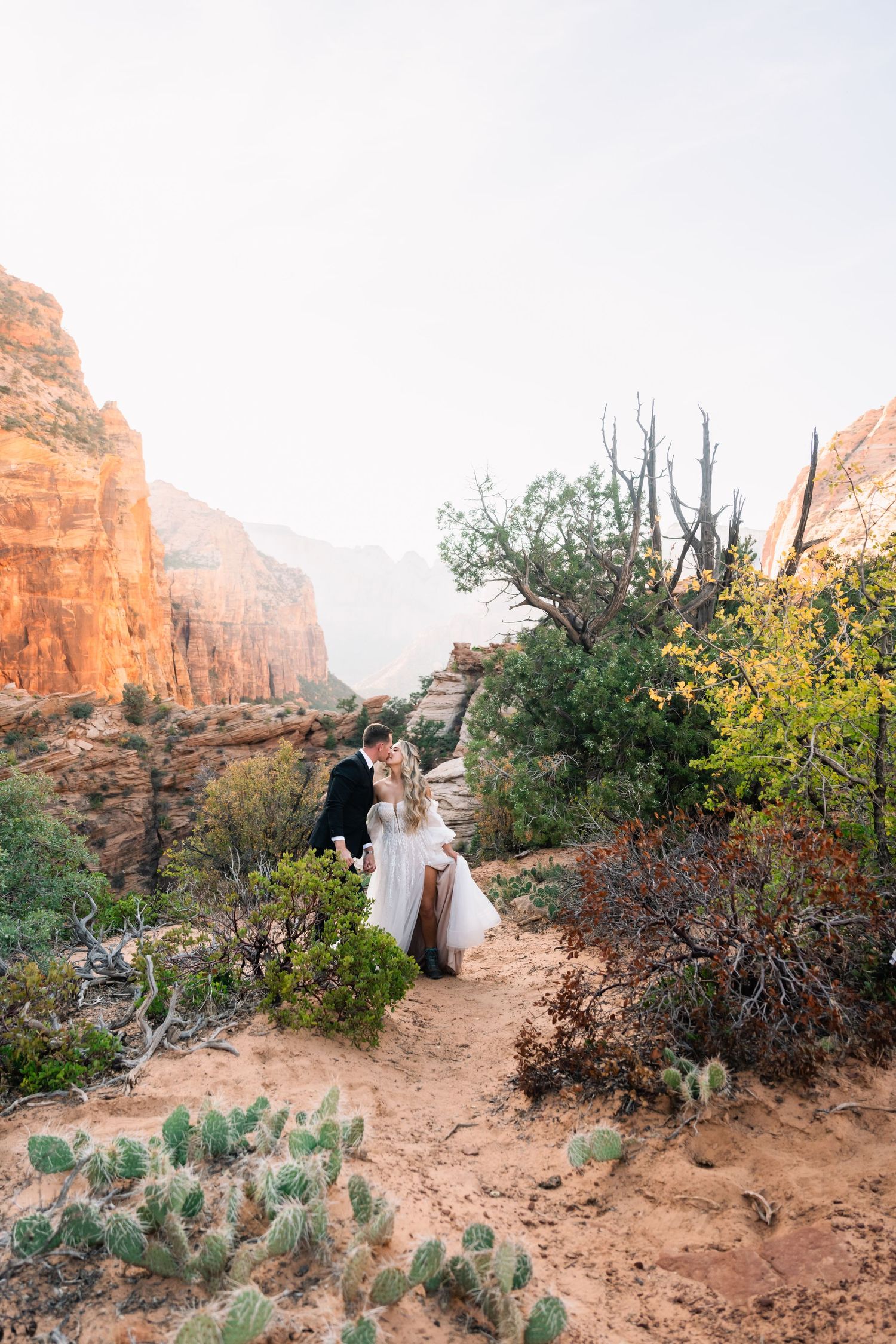 Couple walking desert path surrounded by cacti and red rock formations during wedding photoshoot.