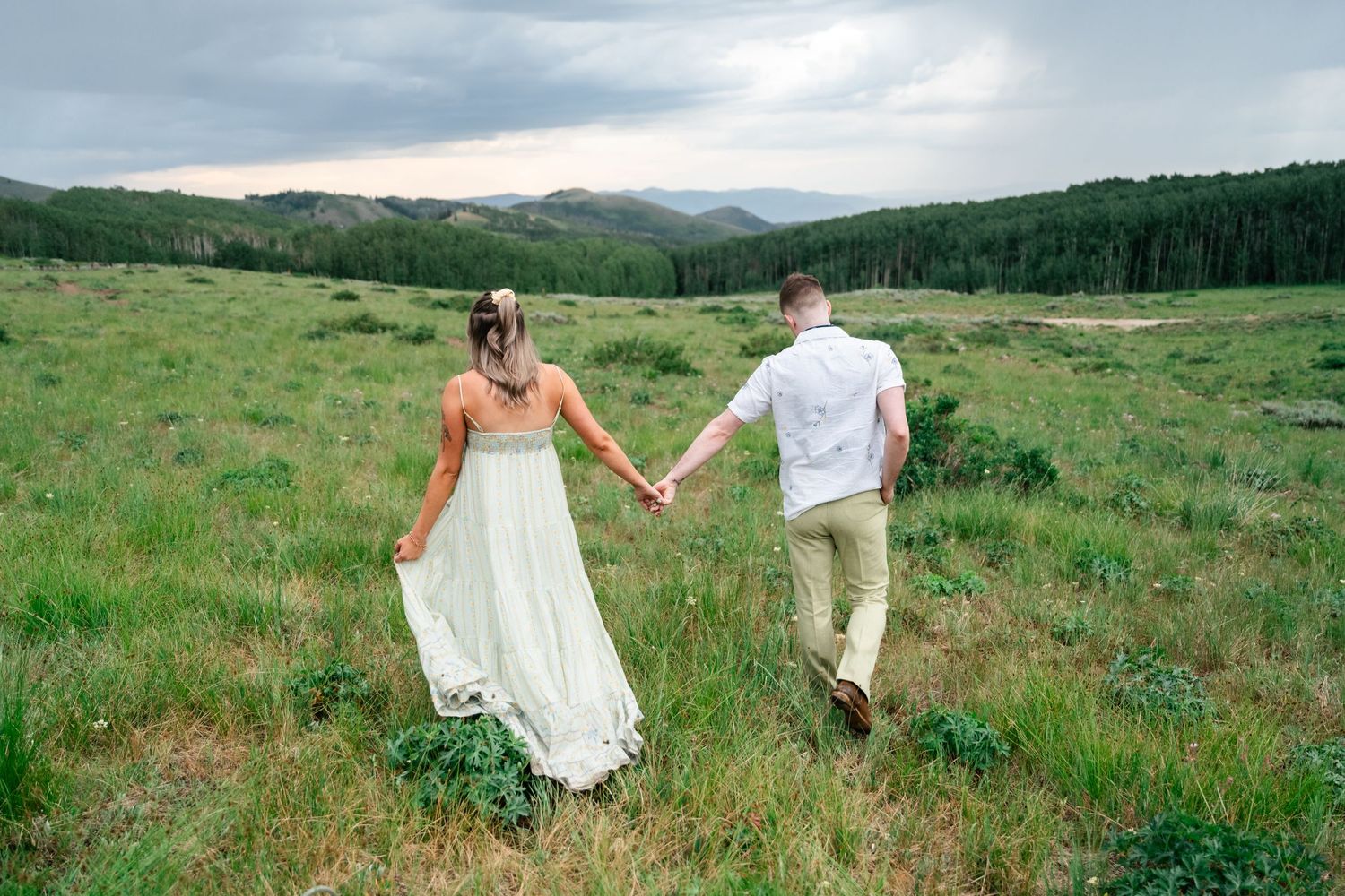 A couple holds hands while walking through a mountain meadow at sunset with rolling hills in the distance. Park City engagement photographer