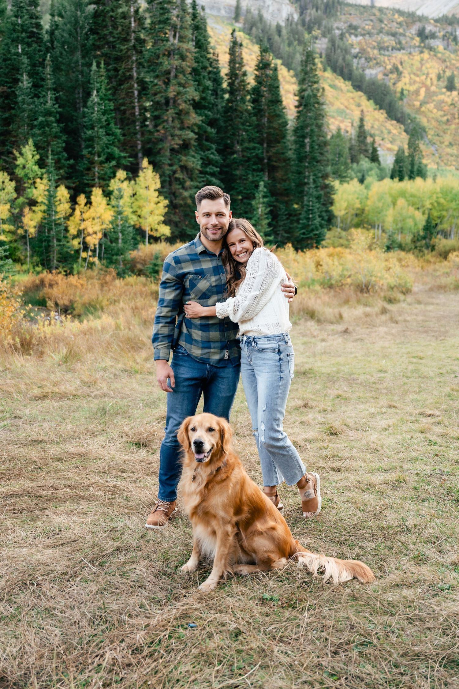 A couple and their Golden Retriever pose together on a mountain trail surrounded by evergreen trees and autumn colors.