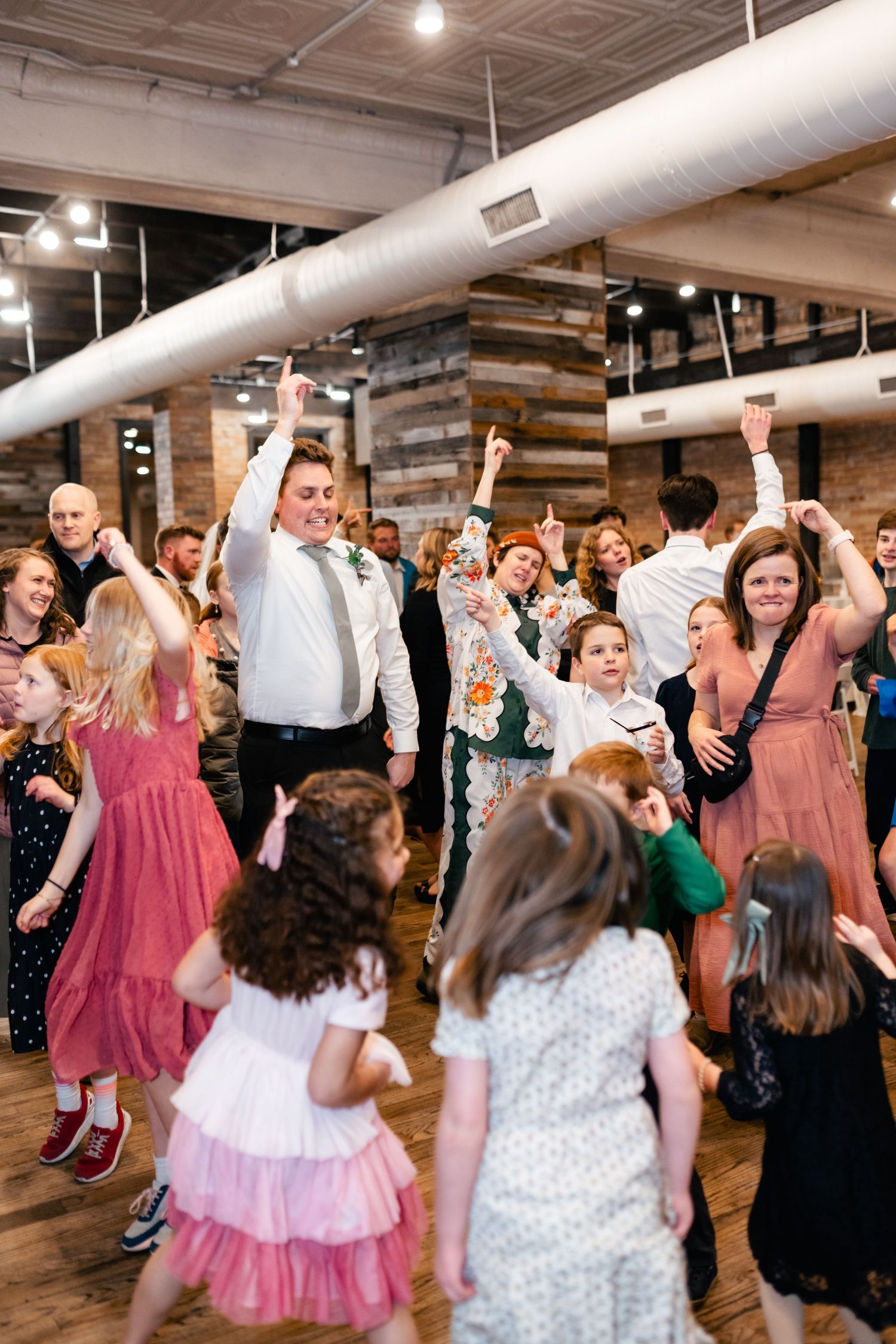 Joyful wedding celebration with guests dancing beneath industrial exposed ductwork and rustic wood accent walls.