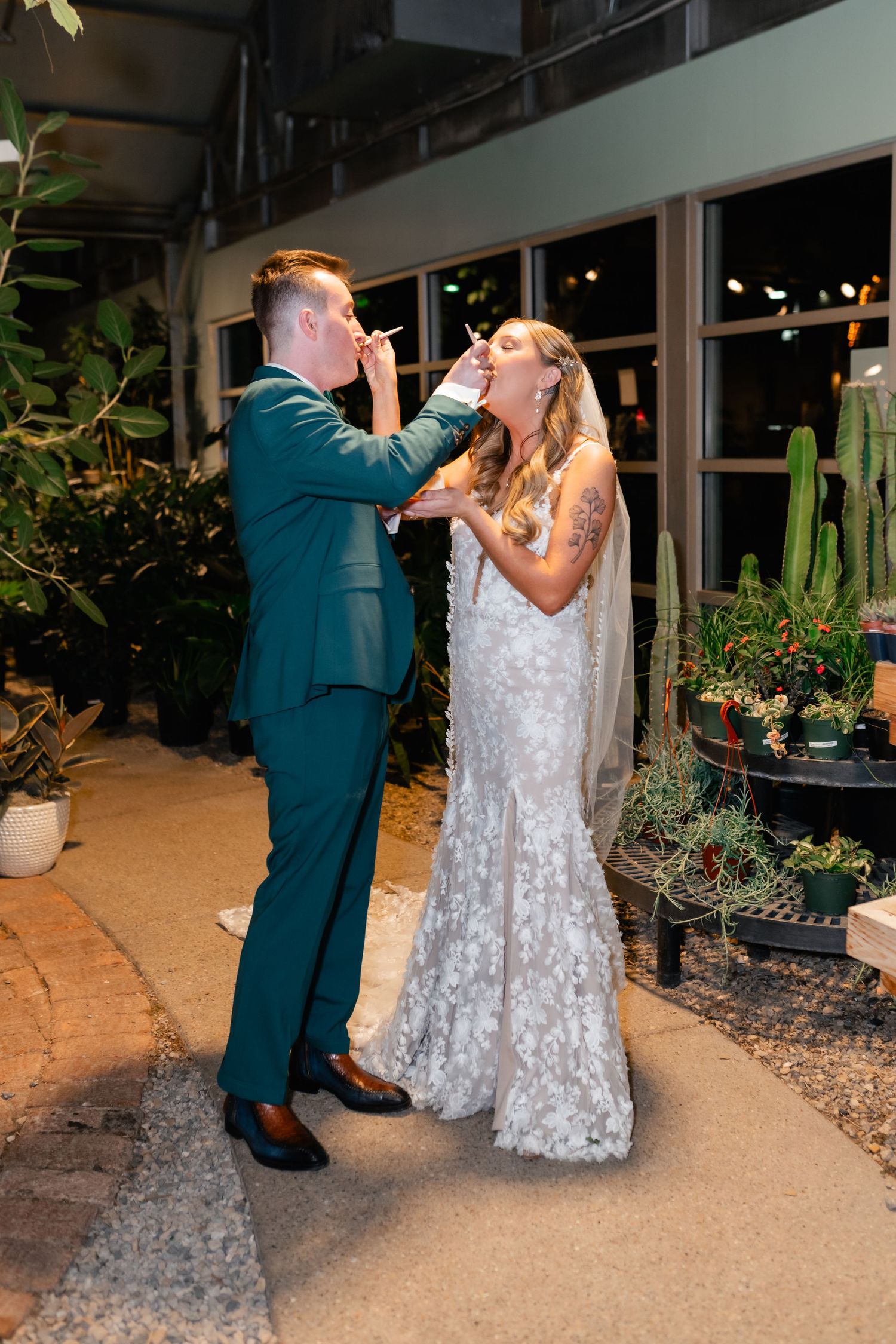 Newlyweds in formal attire playfully share their first bite of wedding cake at an indoor-outdoor reception venue.
