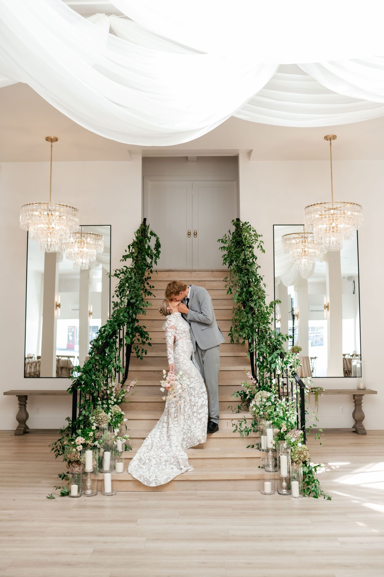 Elegant wedding venue showcases crystal chandeliers, light walls, ornate mirrors, and greenery-wrapped staircase. Utah Wedding Photographer.