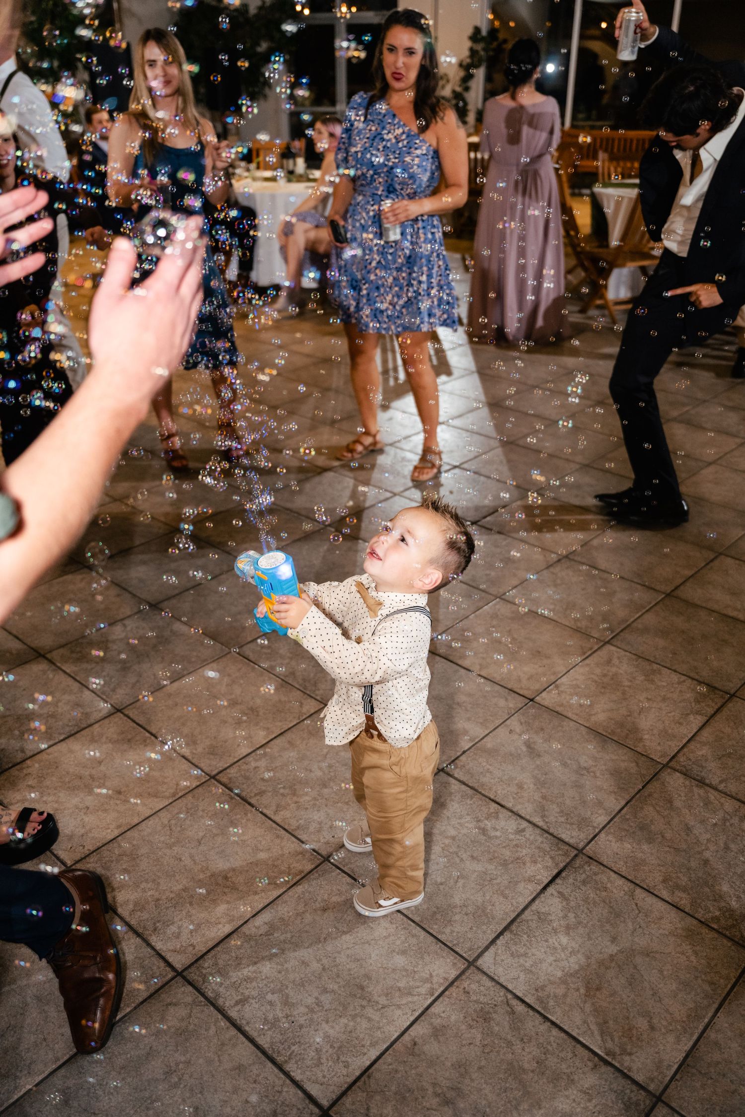 A small toddler in khaki pants plays with bubbles on a tiled dance floor during a celebration.