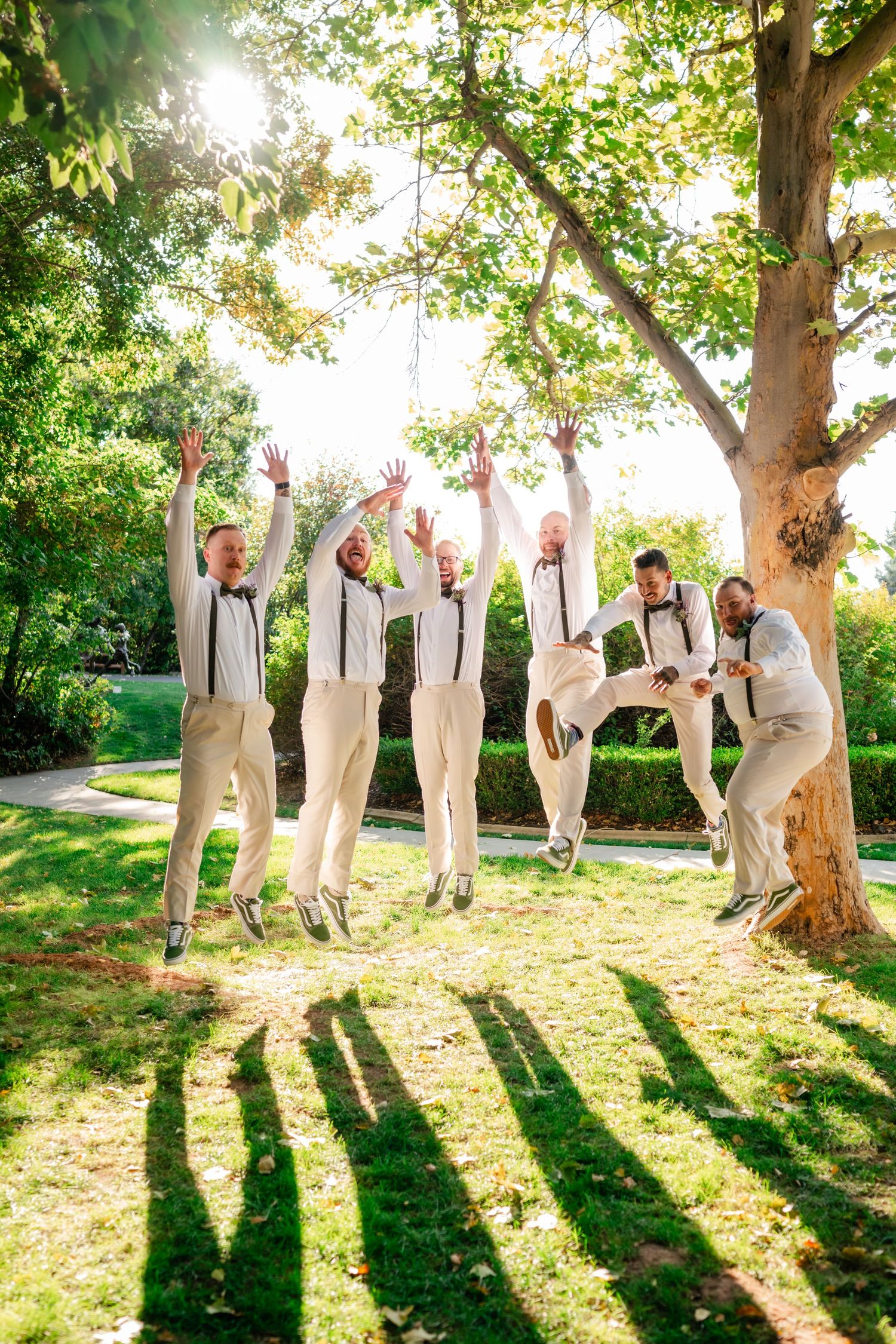 Groomsmen in beige suits and suspenders playfully jumping in unison under a sunlit tree in a garden setting.
