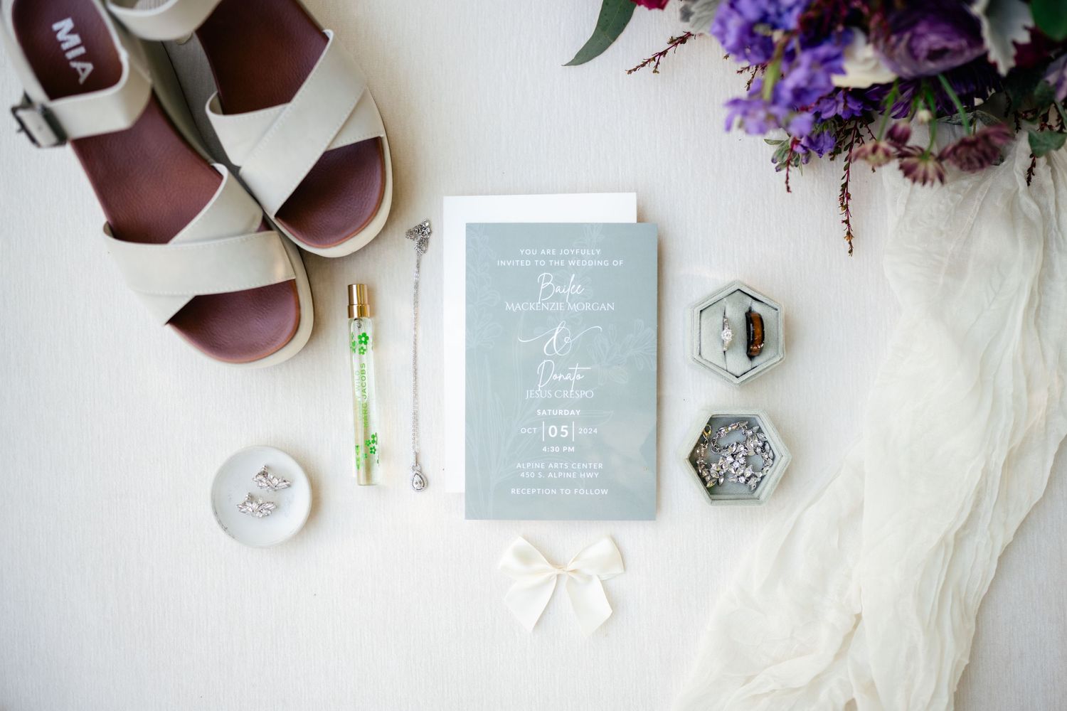 Wedding detail flatlay featuring pale blue invitation, white sandals, purple flowers and decorative accessories on light background.