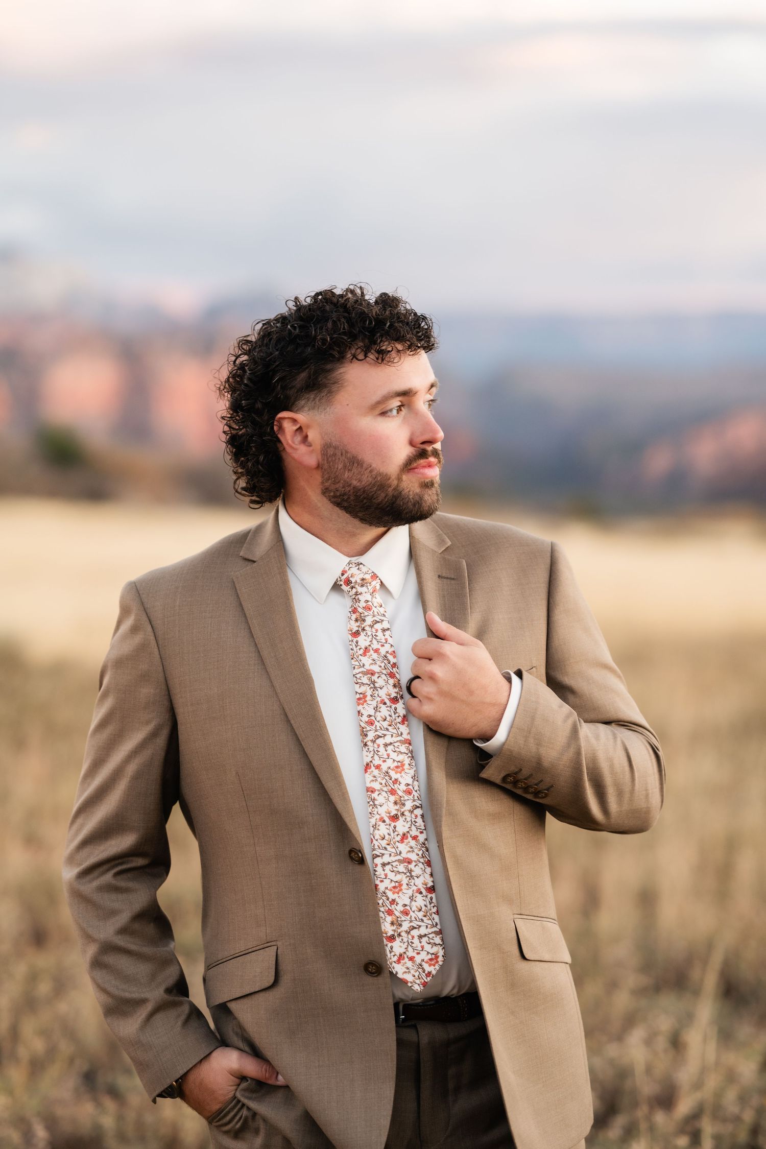 Groom in brown suit and floral tie poses thoughtfully in desert landscape.