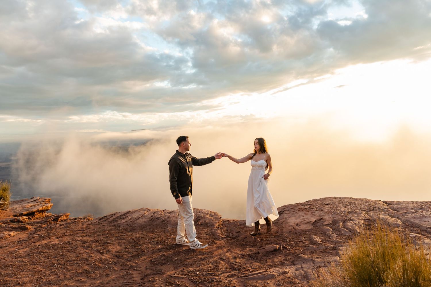 A couple holds hands while standing on a cliff edge during golden hour with clouds below.