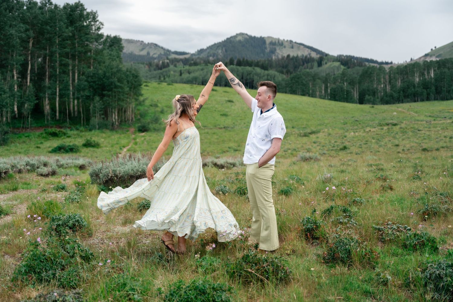 A couple twirls and dances together on a grassy mountainside with dramatic mountain peaks and forest in the distance.