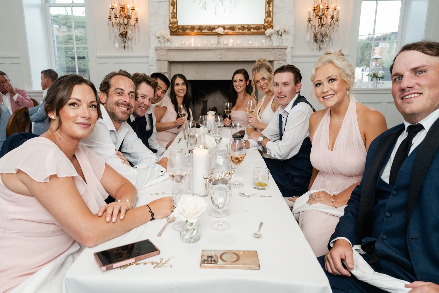 Wedding party seated at an elegant white dining table with crystal chandeliers and gold-framed mirrors.