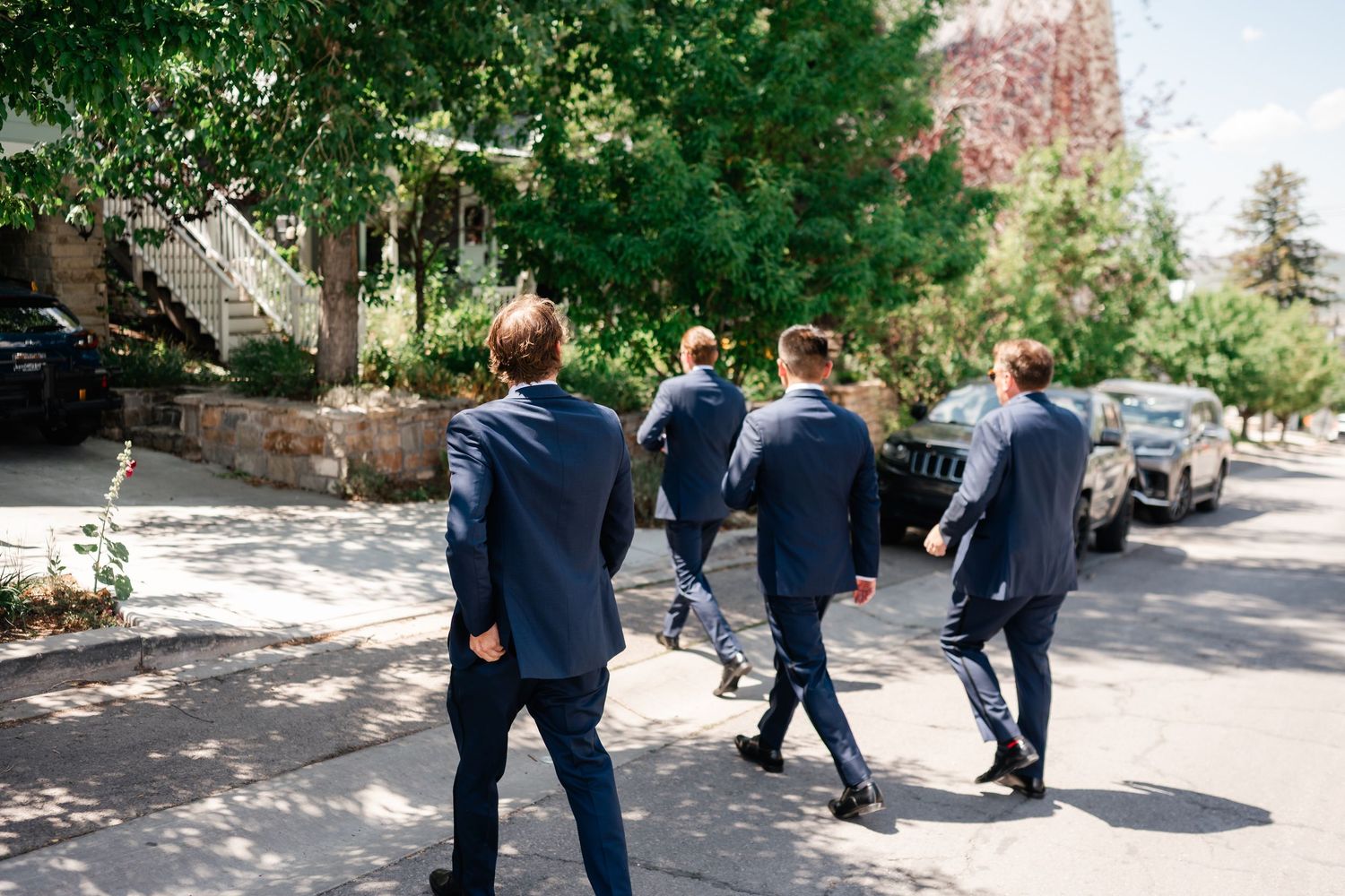Group walking down a tree-lined street wearing navy suits on a sunny day.
