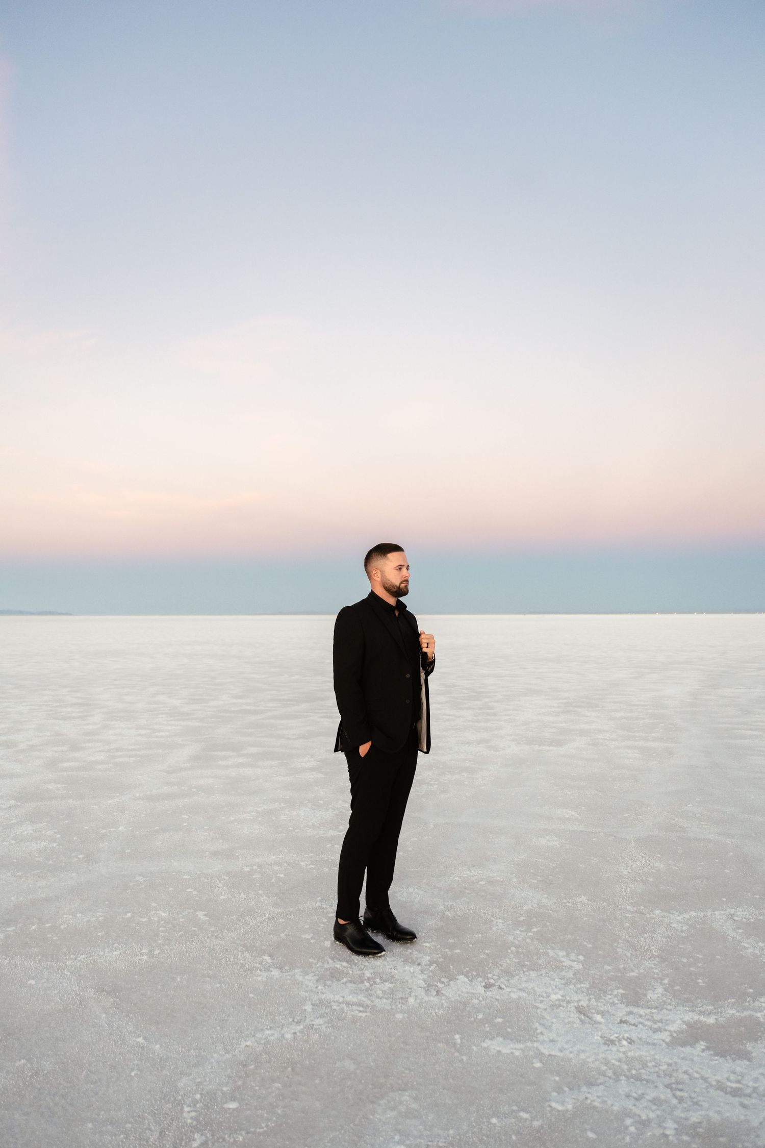 A person in a black suit stands alone on a stark white salt flat landscape at dusk.