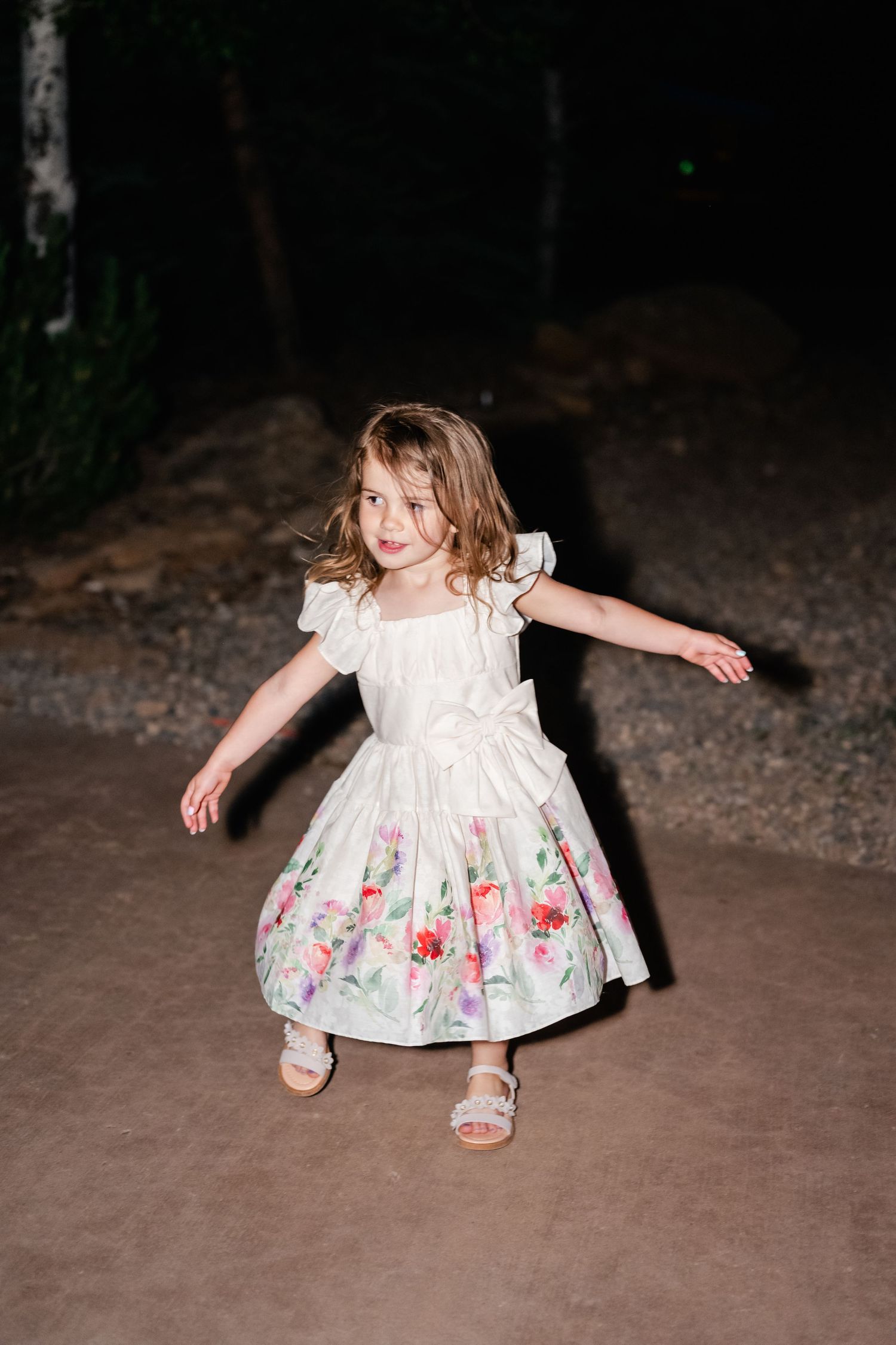 A small figure in a white floral dress twirls and dances on a dirt path at dusk.