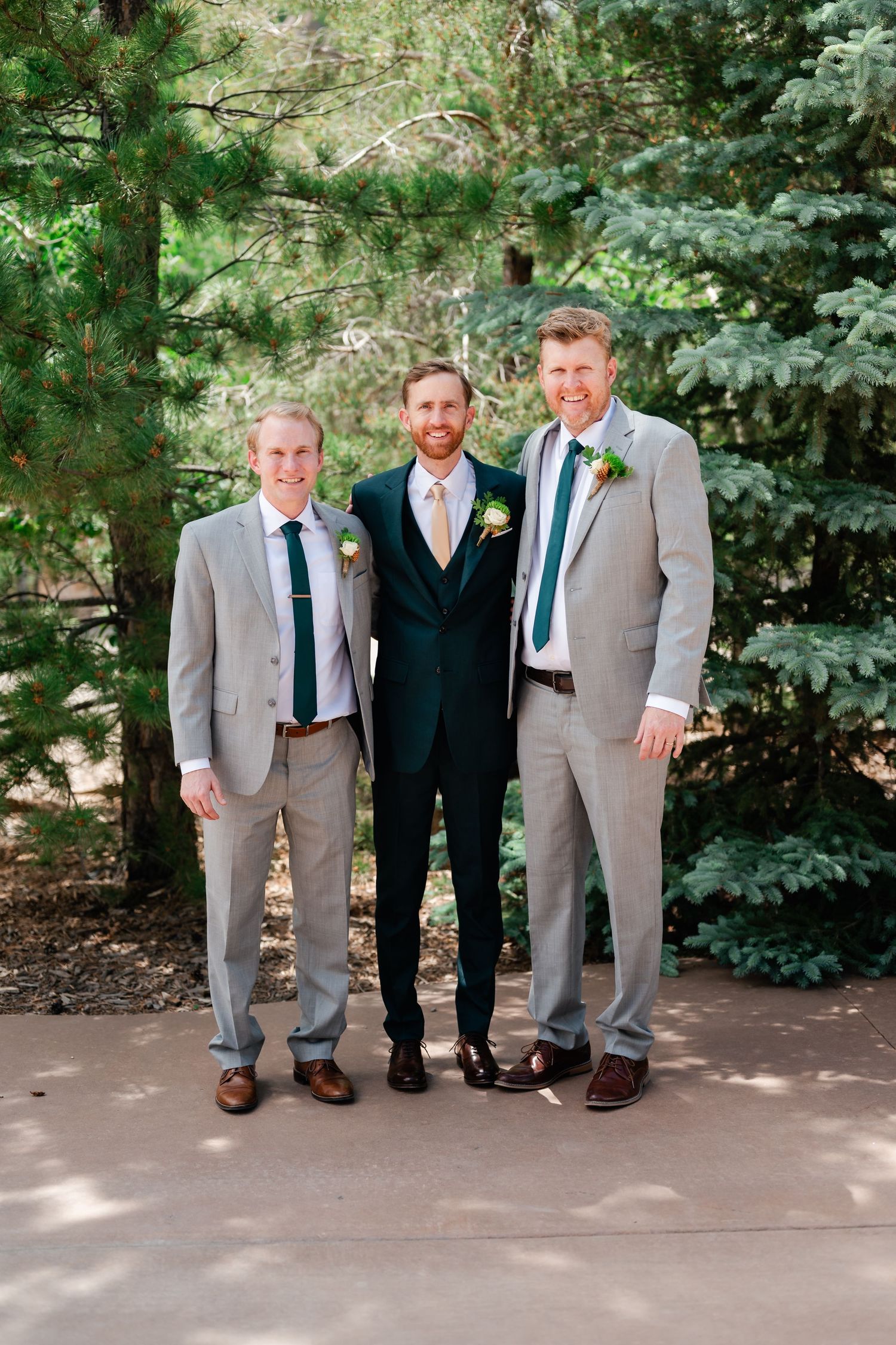 Three men in suits pose together against evergreen trees.