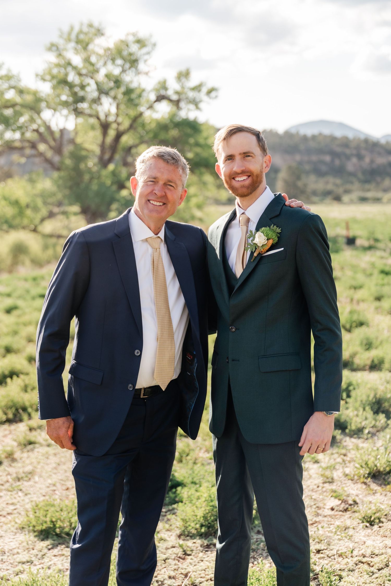 Two men in dark suits and gold ties share candid moments while standing in a desert landscape with mountains in background.