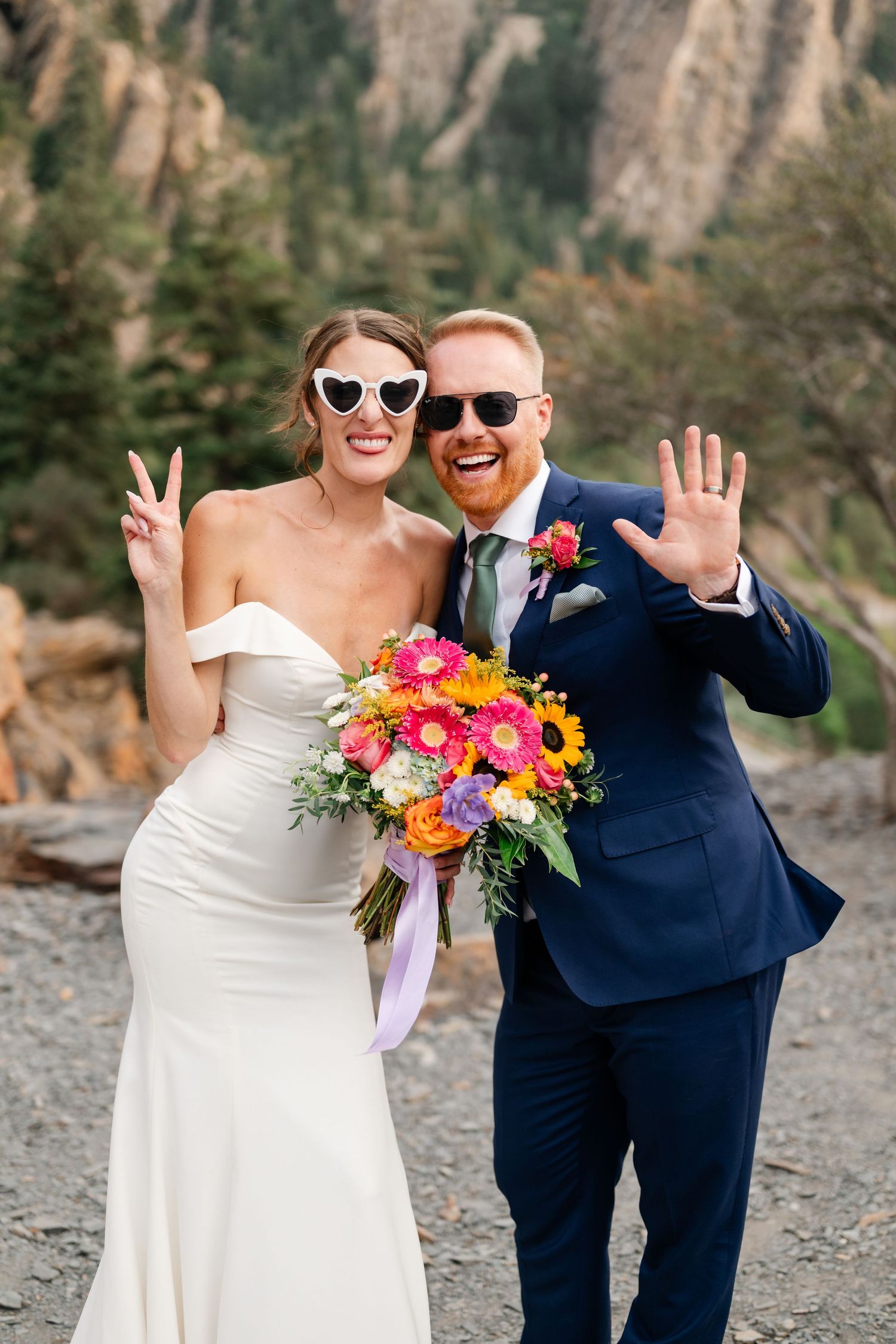 Bride and groom in sunglasses smiling and waving outdoors with colorful bouquet and mountain backdrop.