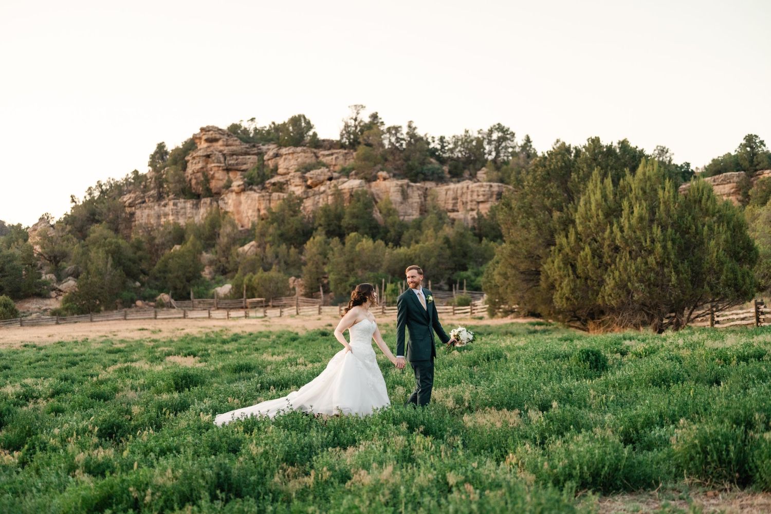 Wedding couple strolls through tall grass with mountain landscape behind them.