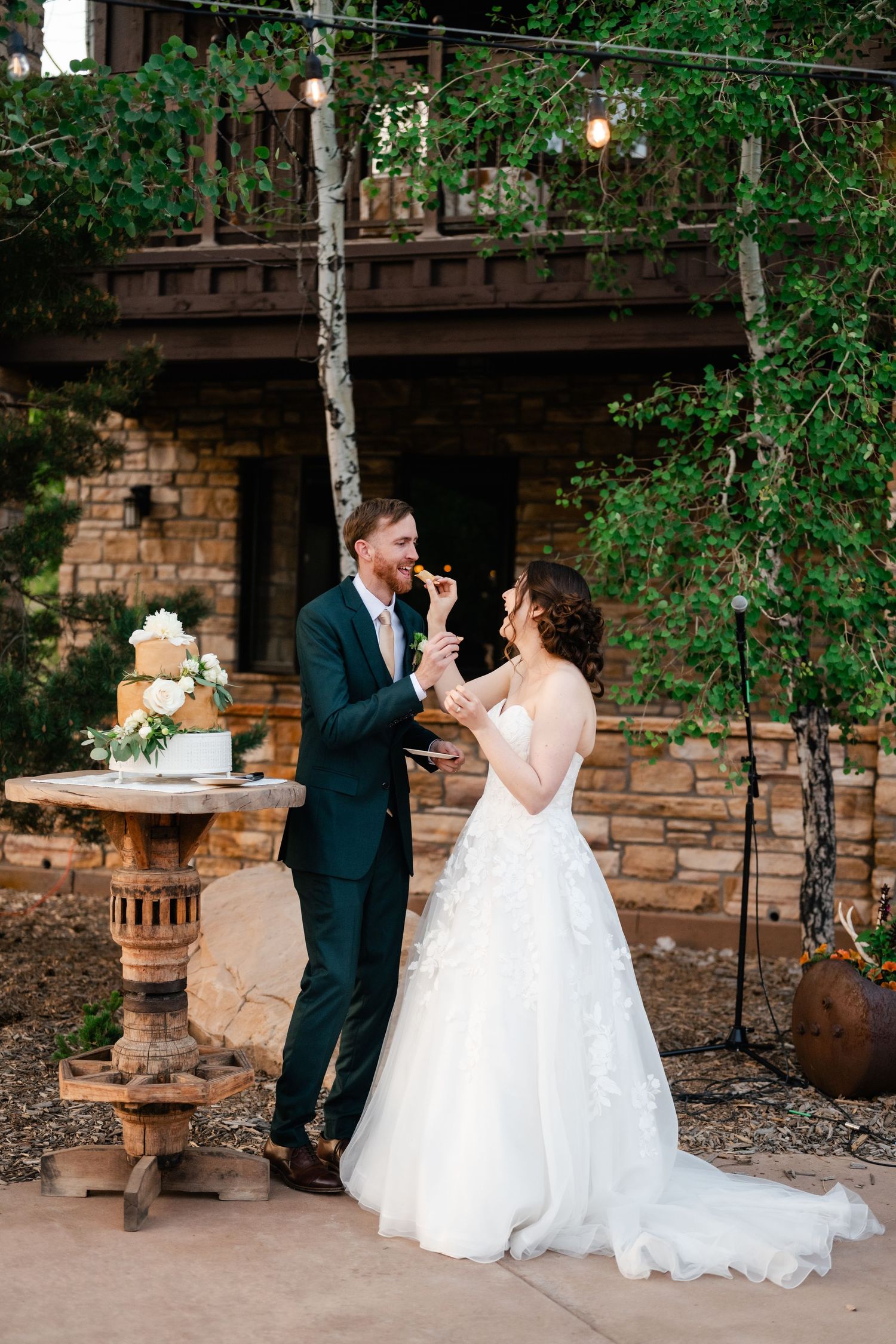 A couple shares their first bite of wedding cake together in an outdoor garden setting with stone walls and greenery.