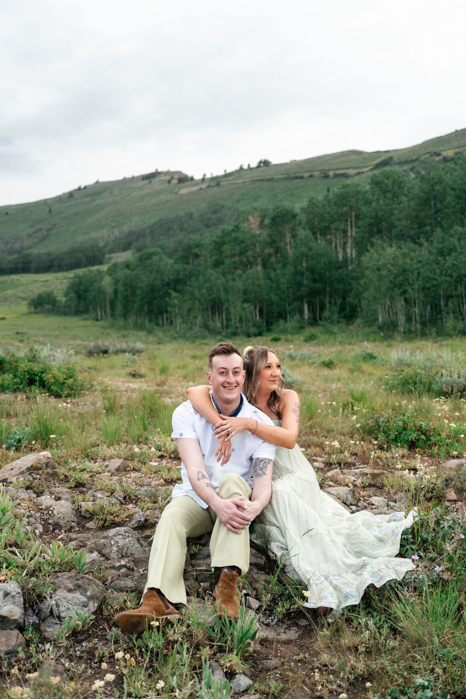 A romantic couple relaxes on rocky ground with mountains and forest creating a scenic backdrop.