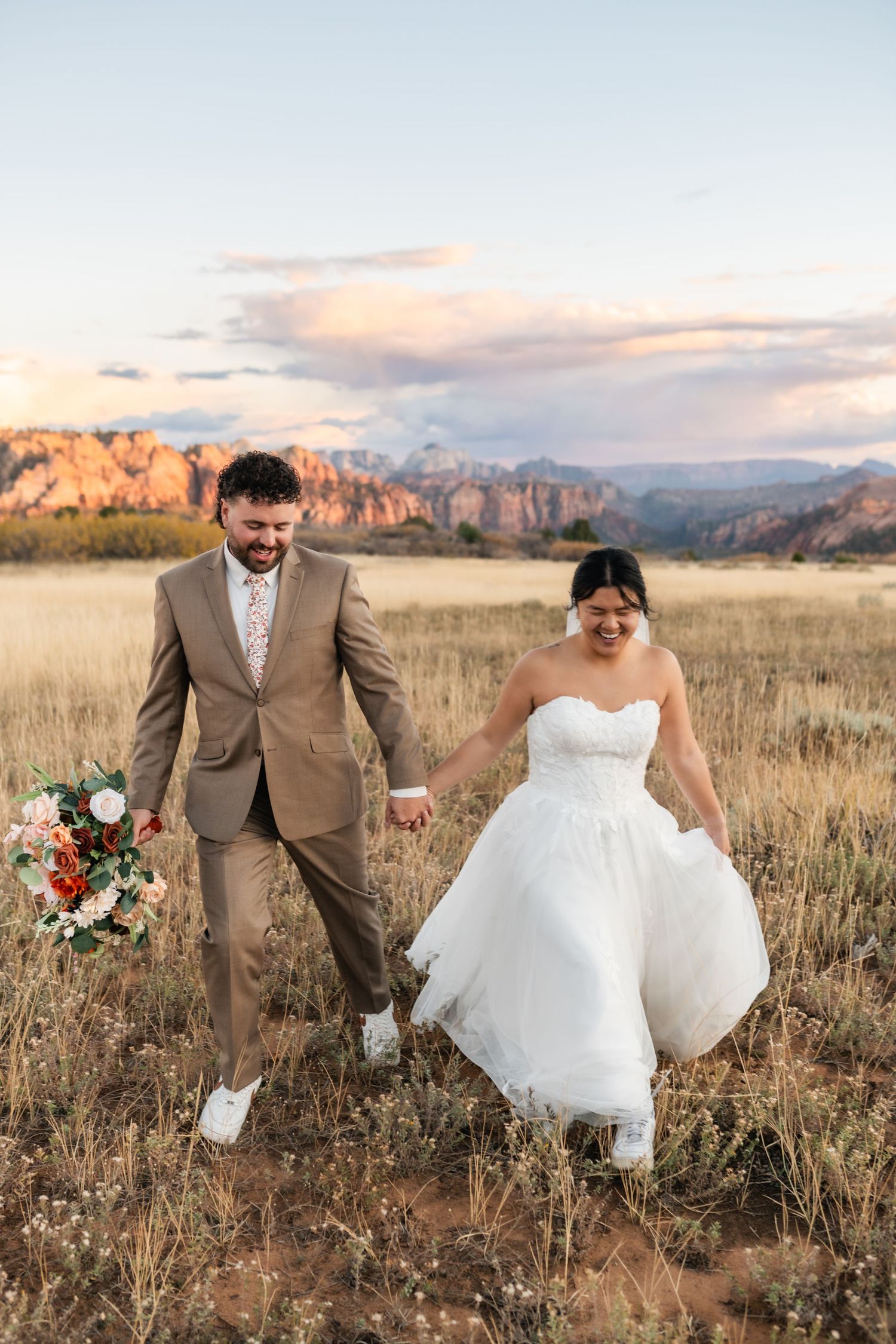 A couple joyfully walks through a field with mountains and sunset in the background.