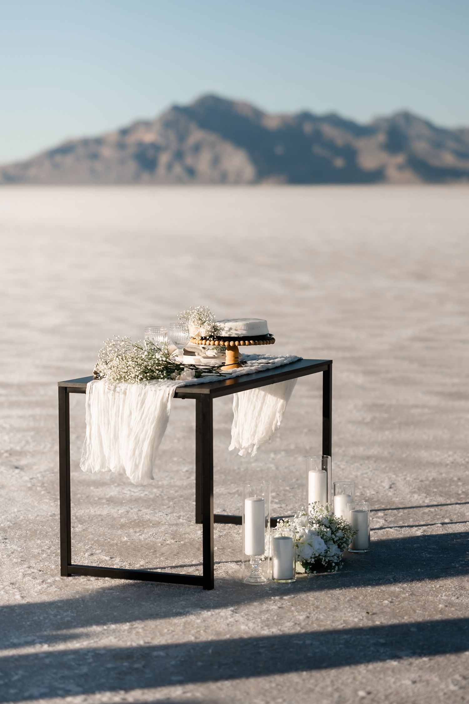 Modern black tables with white decor and flowers set up on salt flats with mountains in background.