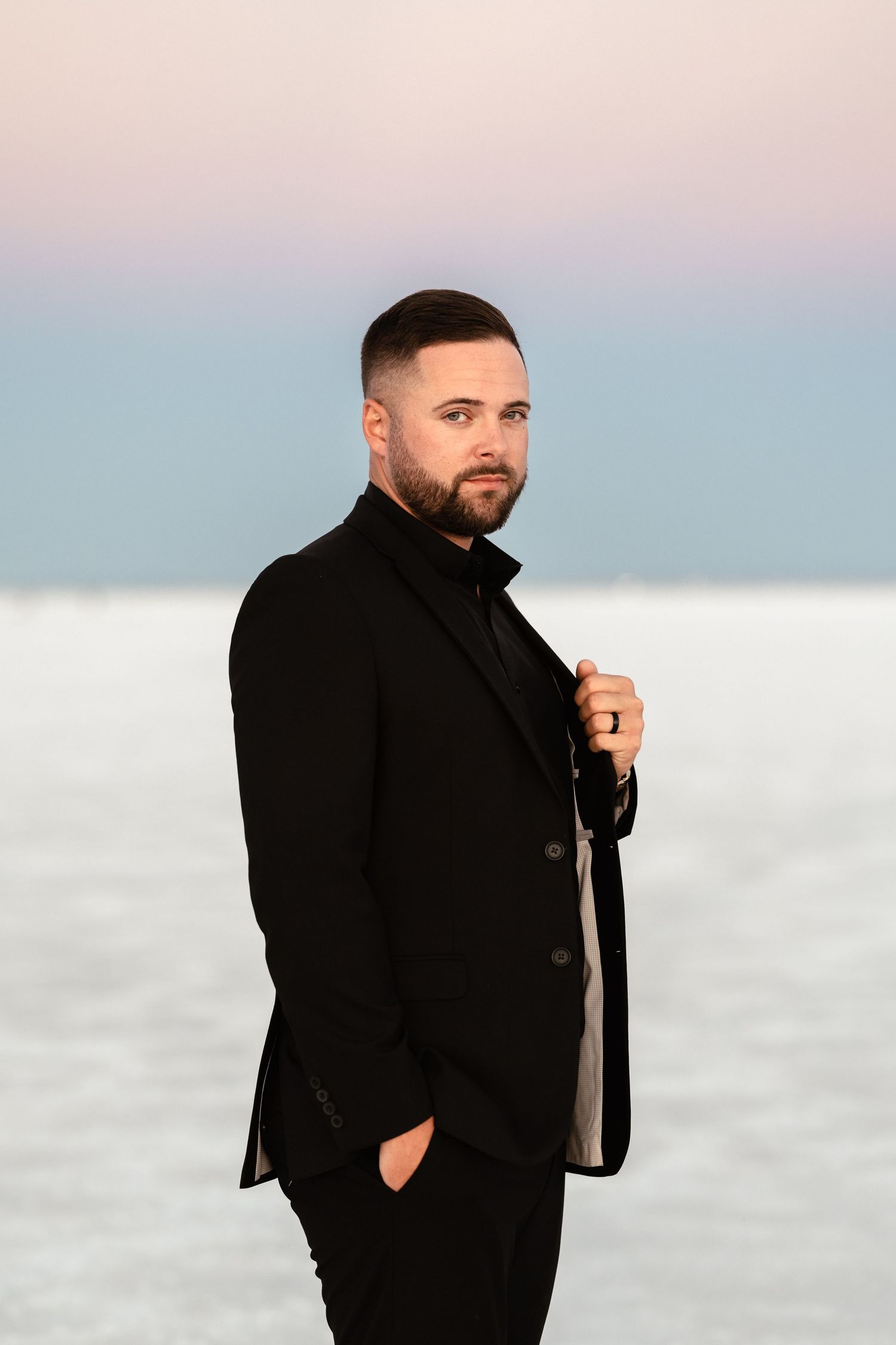 Groom in black suit poses against pastel sky and white salt flats landscape.