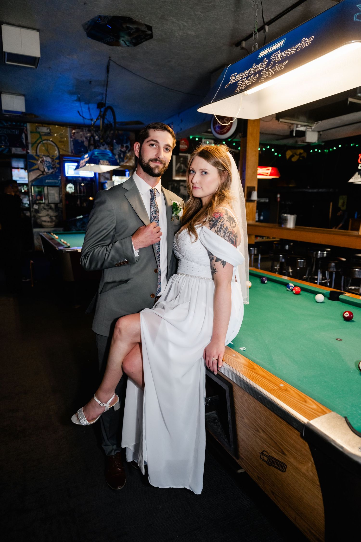 A bride and groom strike a fun pose together against a pool table in a dimly lit bar setting.