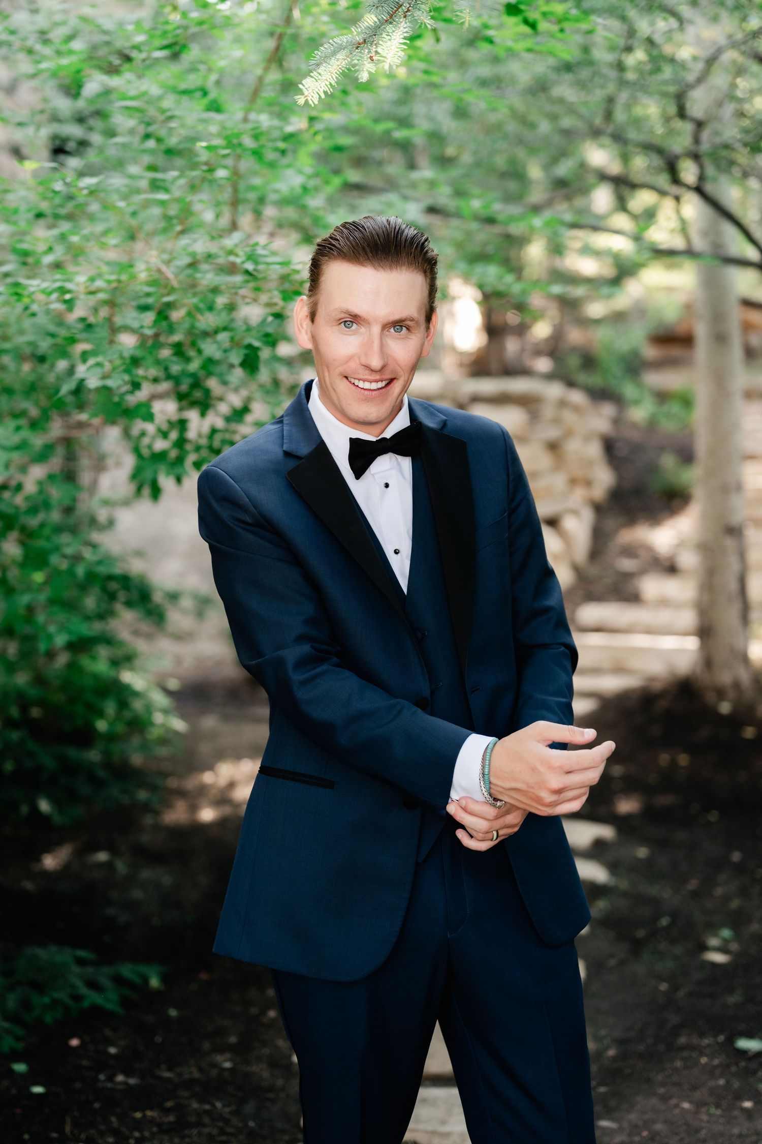 A groom in a classic black tuxedo smiles while adjusting his cuff in a garden setting.