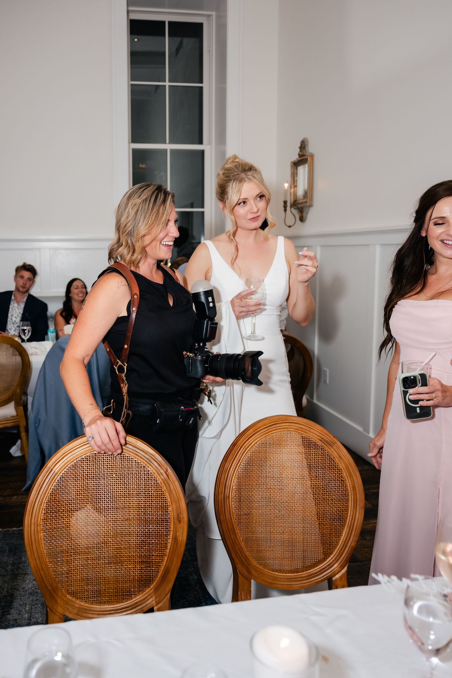 Wedding reception guests gather around elegant wooden chairs in an indoor venue with white tablecloths.