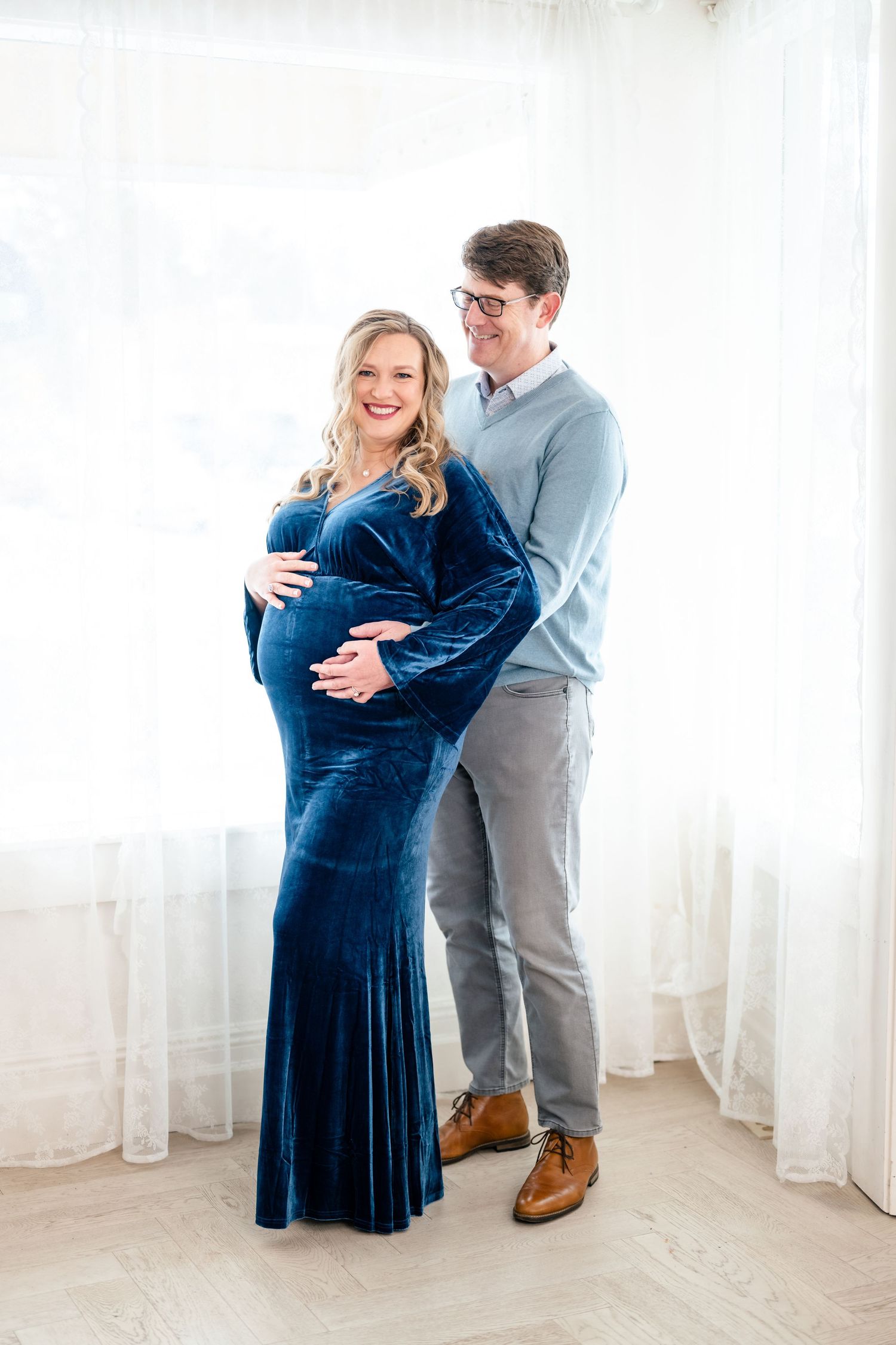 A couple in a light-filled room shares tender moments as they pose for maternity photos in blue velvet dress and gray sweater.