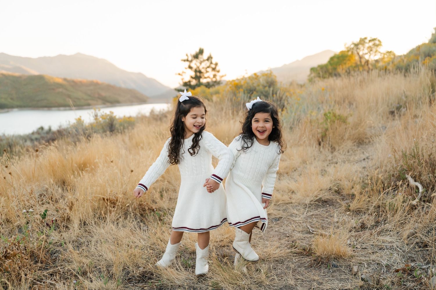 Two people in white outfits walk through a golden field at sunset in the mountains.