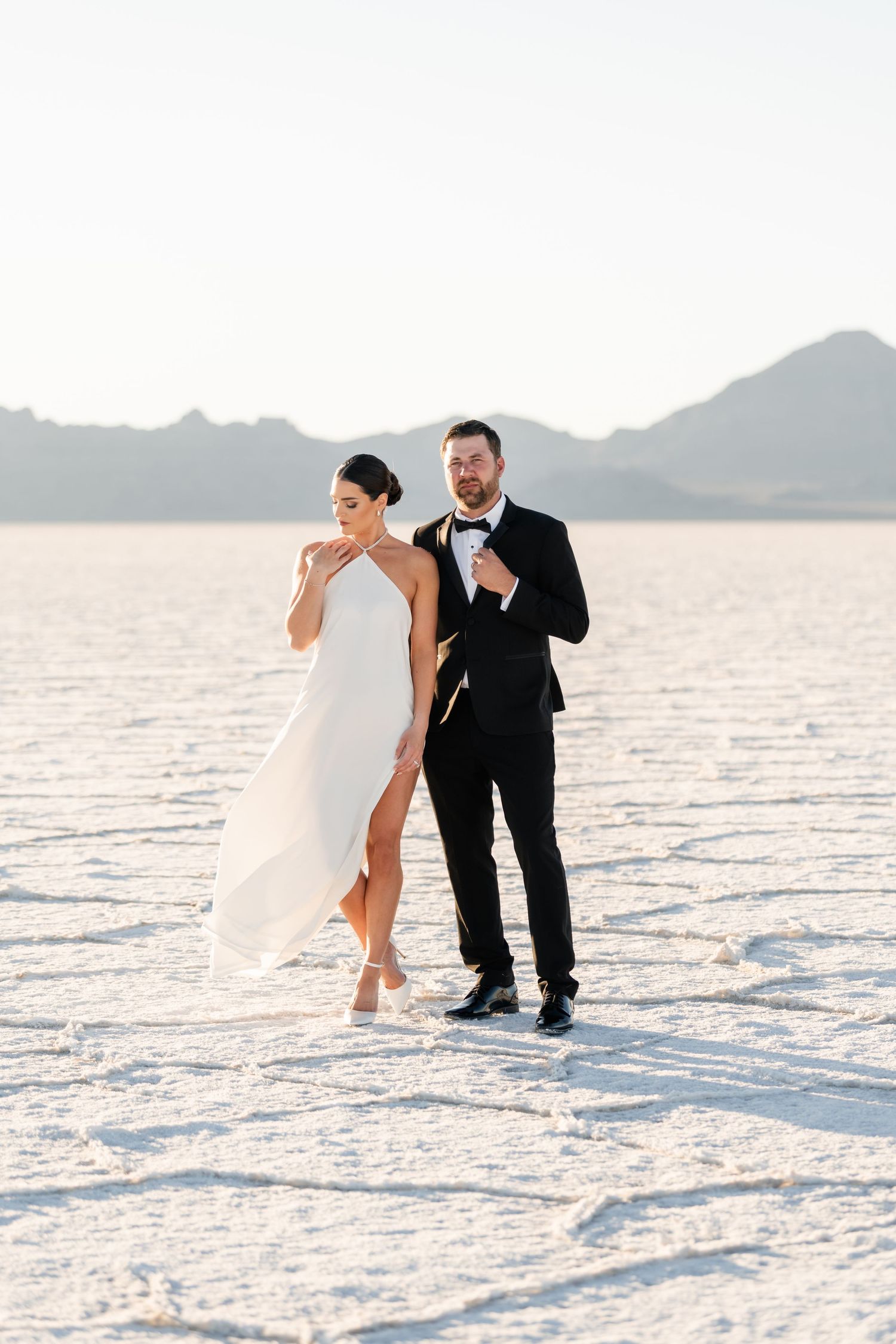A couple in wedding attire poses playfully on the white salt flats with mountains in the background. Salt Flats Photographer.