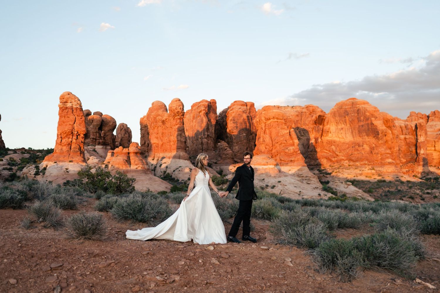 A couple in wedding attire stands silhouetted against stunning red rock formations at Arches National Park during sunset.