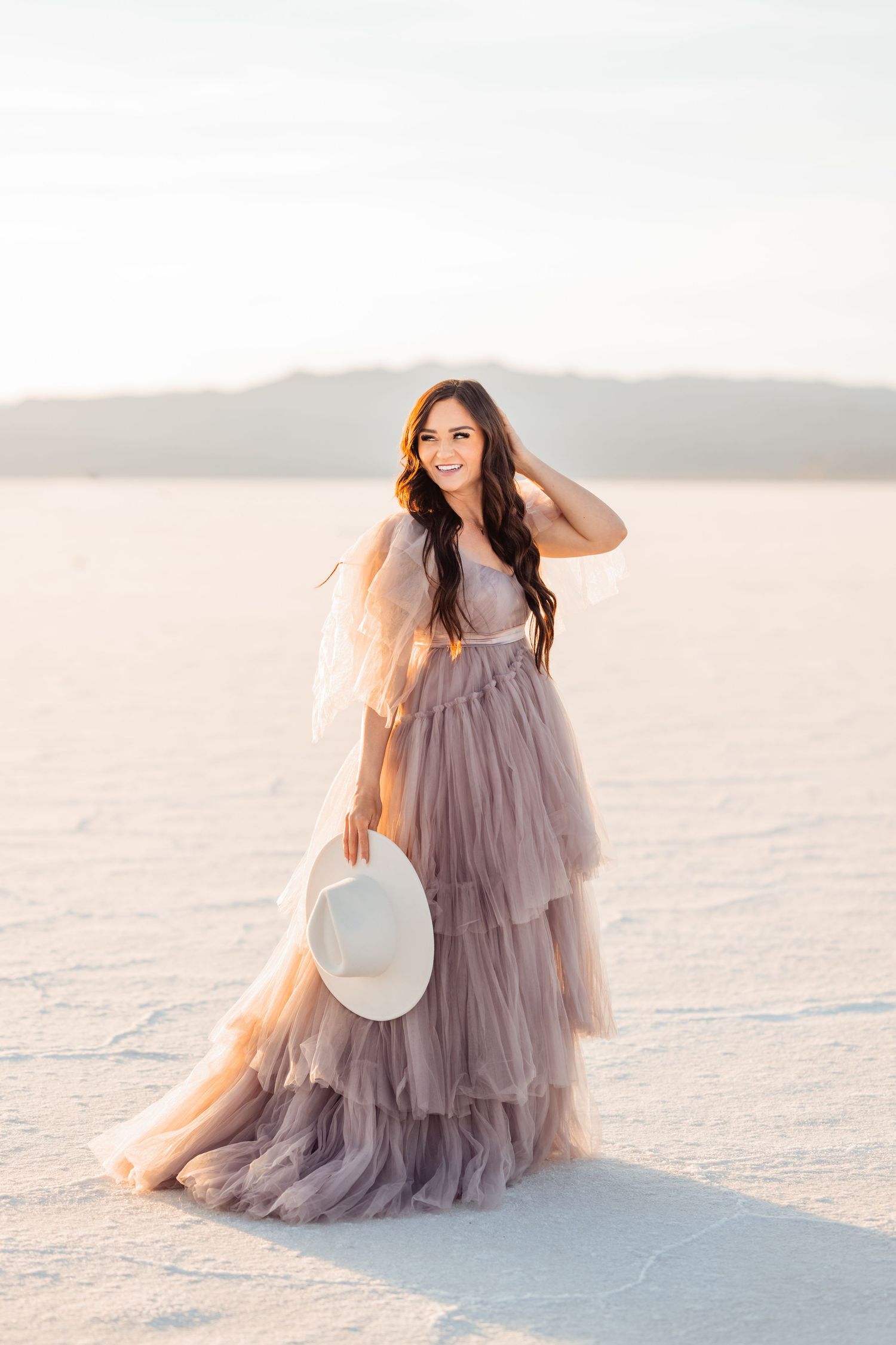 Utah Salt Flats Photographer. Woman in flowing lavender tulle gown holds white hat while posing on salt flats at sunset.