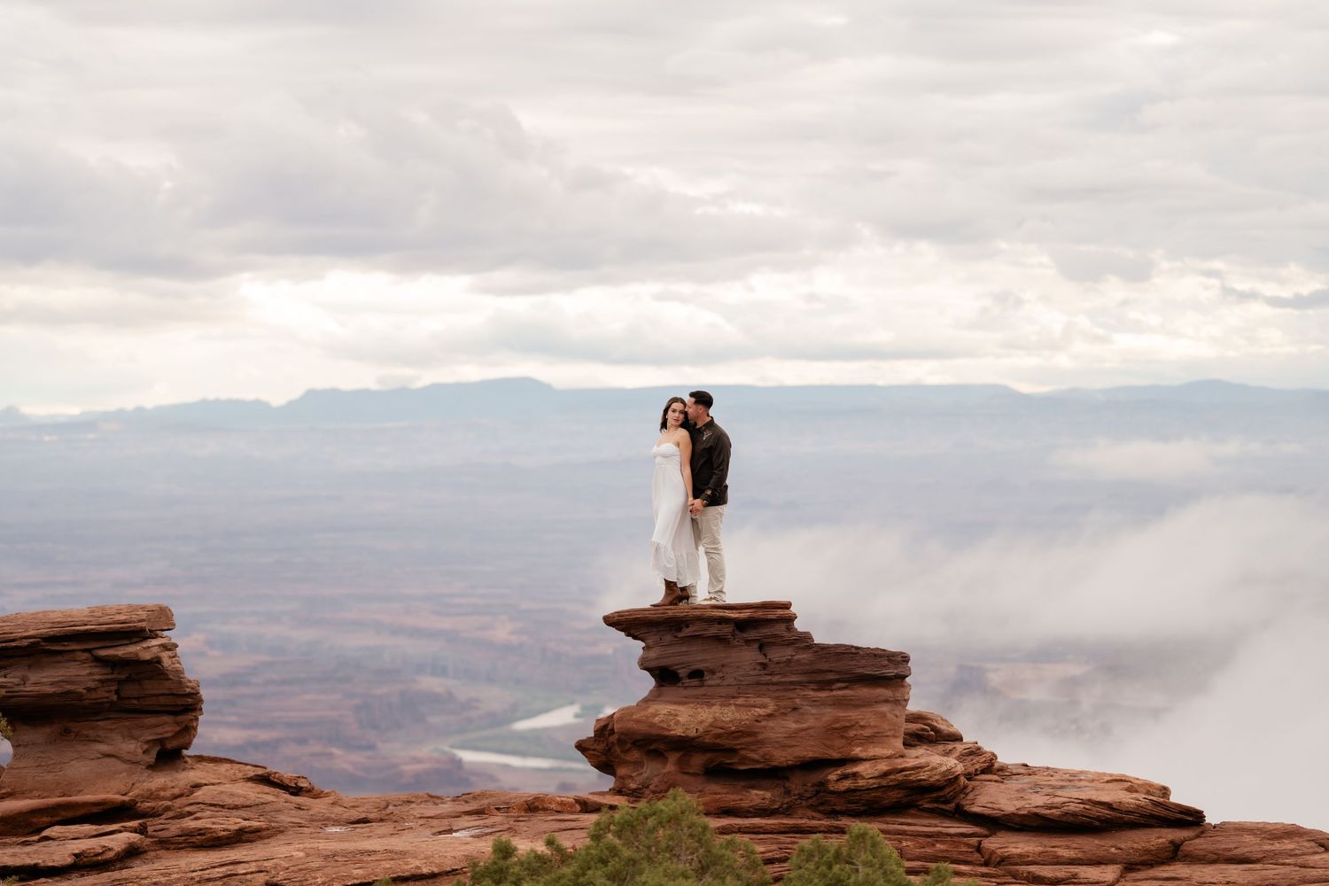 Silhouetted figures pose on a scenic red sandstone cliff edge against a cloudy mountain backdrop.
