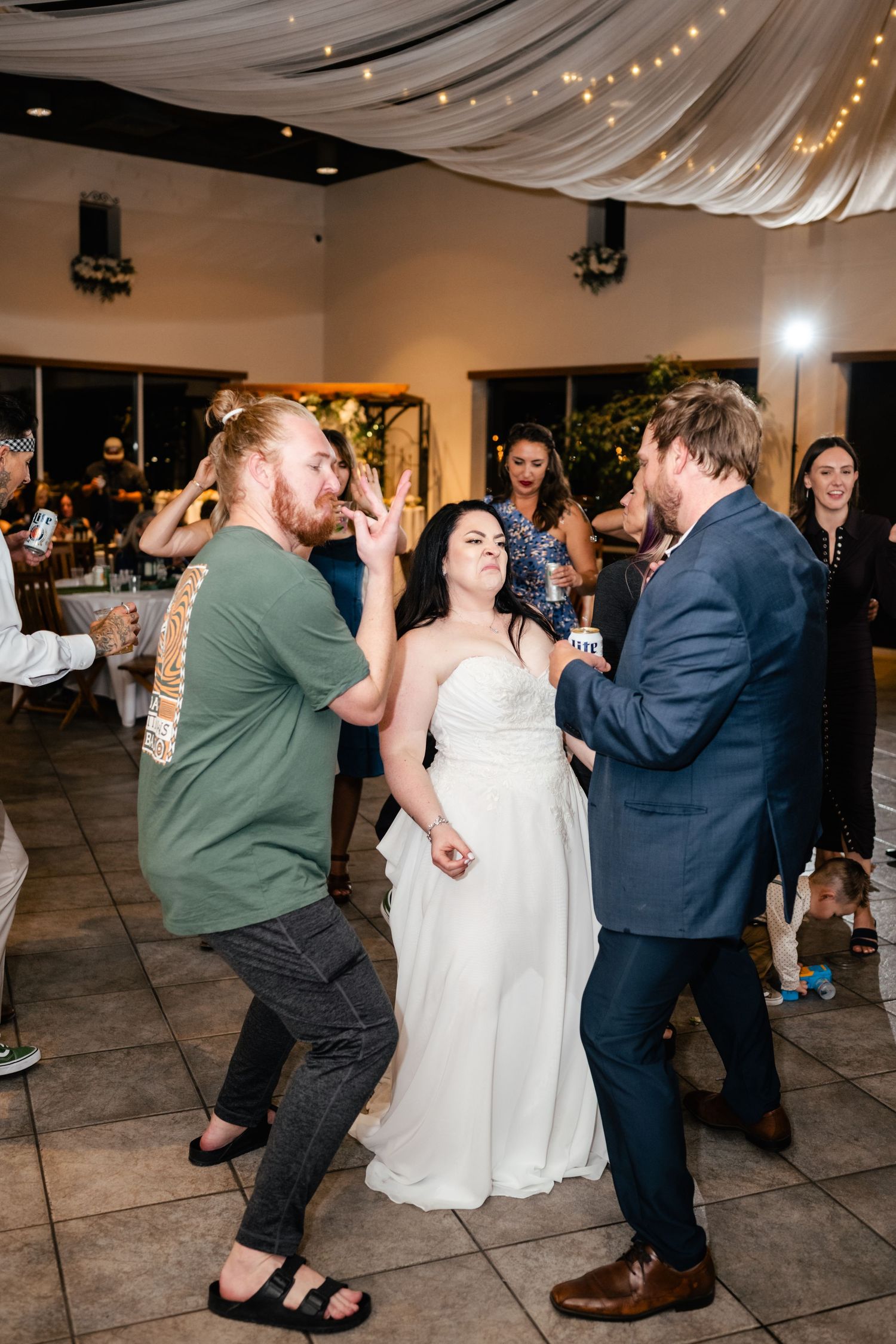Three wedding guests dance together at a reception under draped fabric and string lights in an indoor venue.