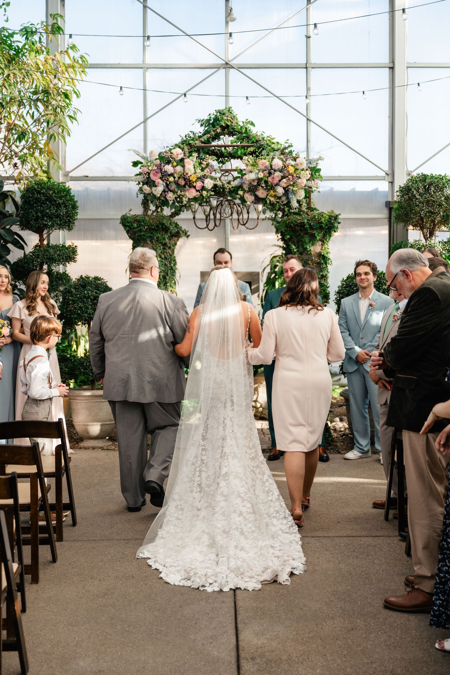 Wedding ceremony procession in a greenhouse with floral arch and long lace veil trailing behind.