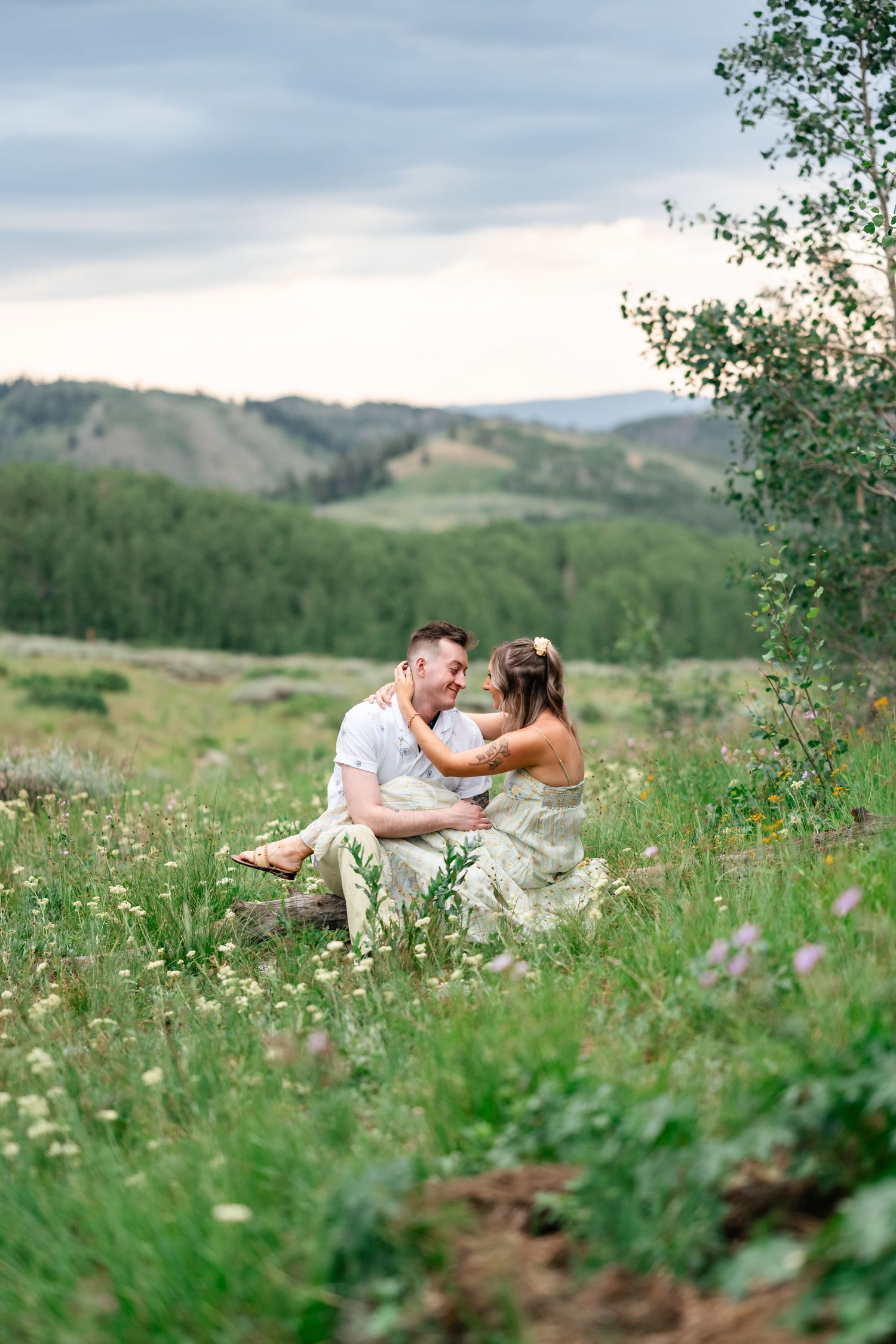 A couple embraces while sitting in a wildflower meadow with rolling hills and mountains in the background. Utah mountain engagement session.