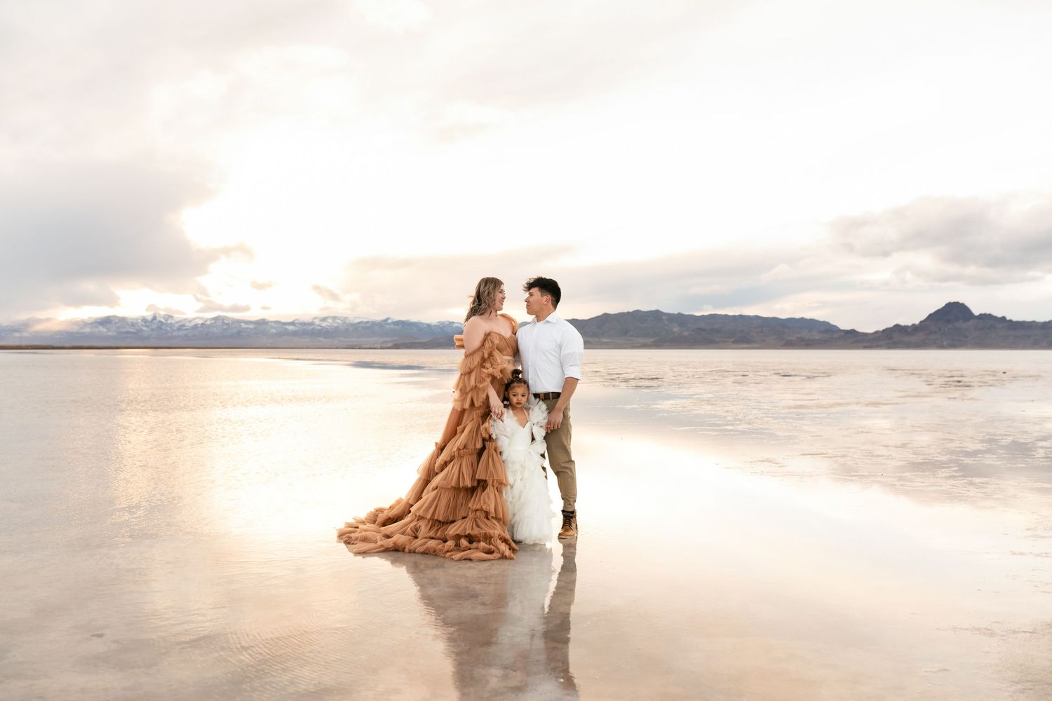 A couple in wedding attire standing on reflective salt flats at sunset with mountains in the distance.