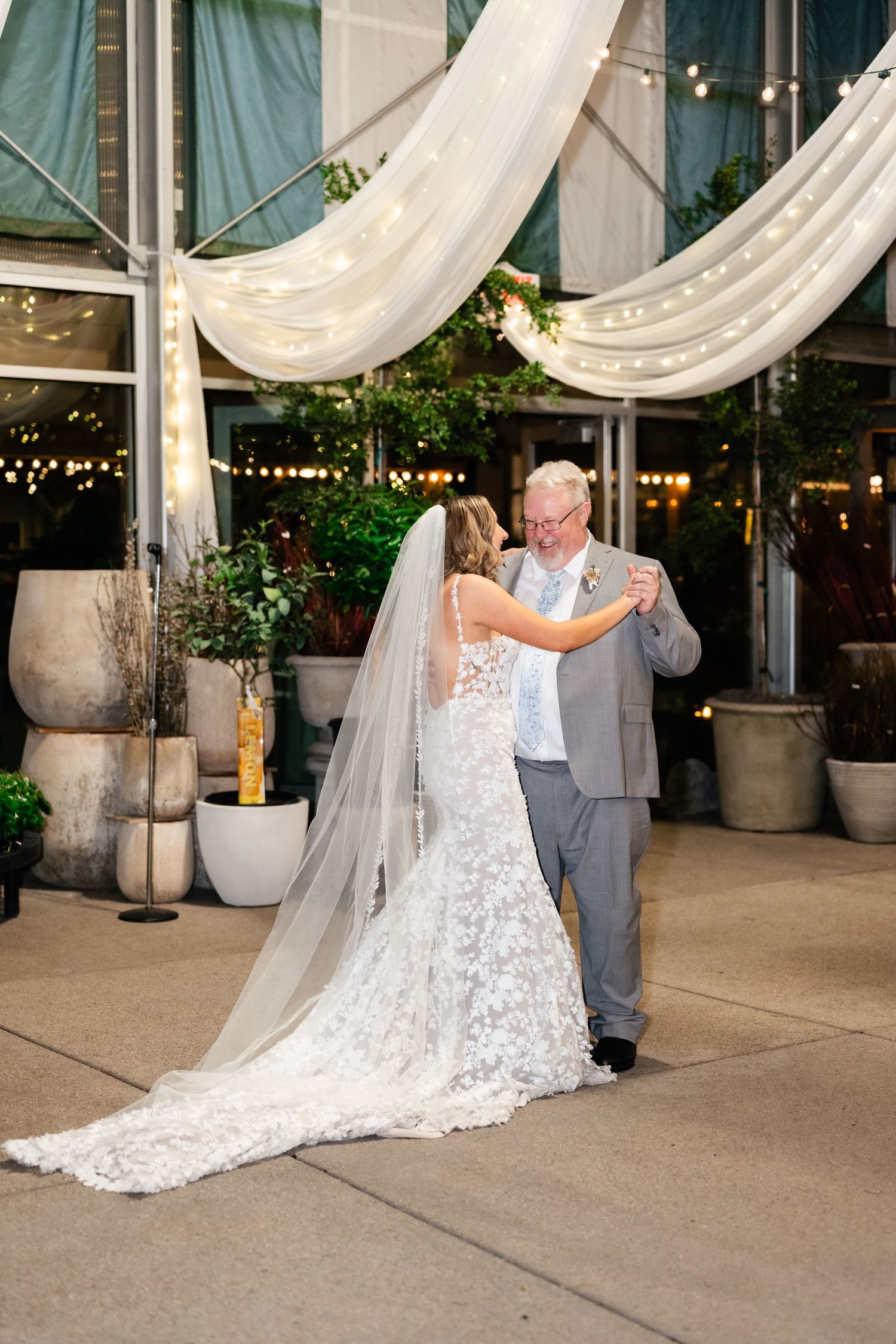 A bride in a lace wedding dress with long veil dances with someone in a gray suit under elegant white draping and string lights.