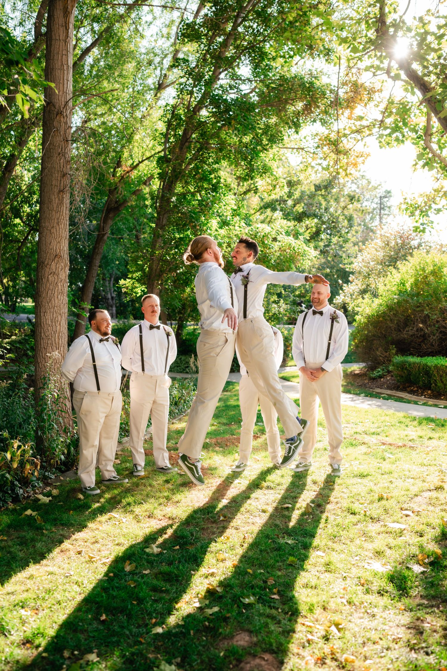 Wedding party in light colored attire playfully poses beneath tall trees casting long shadows in afternoon sunlight.